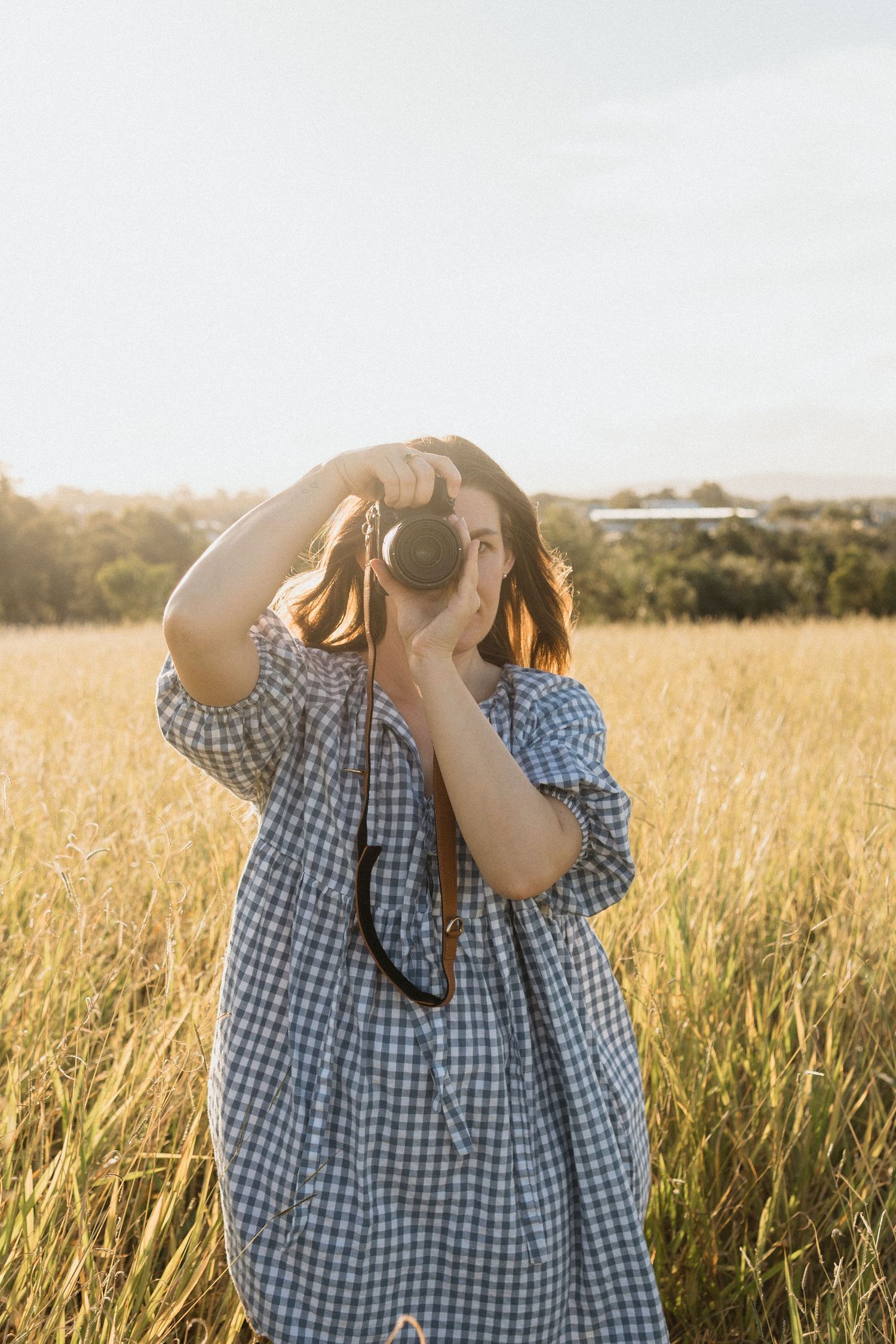 Woman in a blue gingham dress walking through a field and capturing photos with a camera.