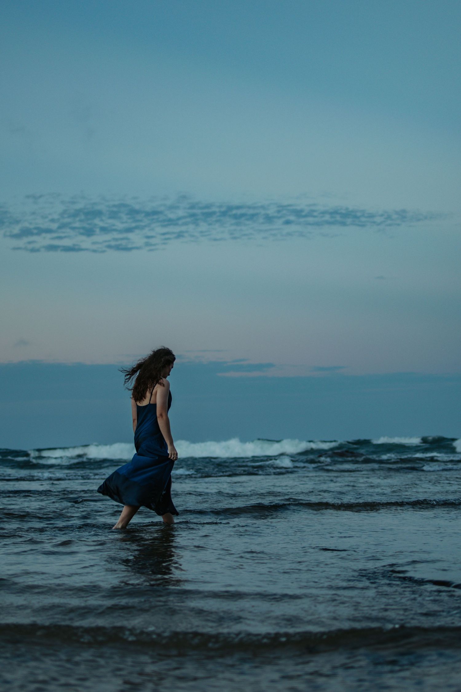 A woman in a blue dress wades through dark ocean waters at dusk with waves crashing against rocky shores in the background.