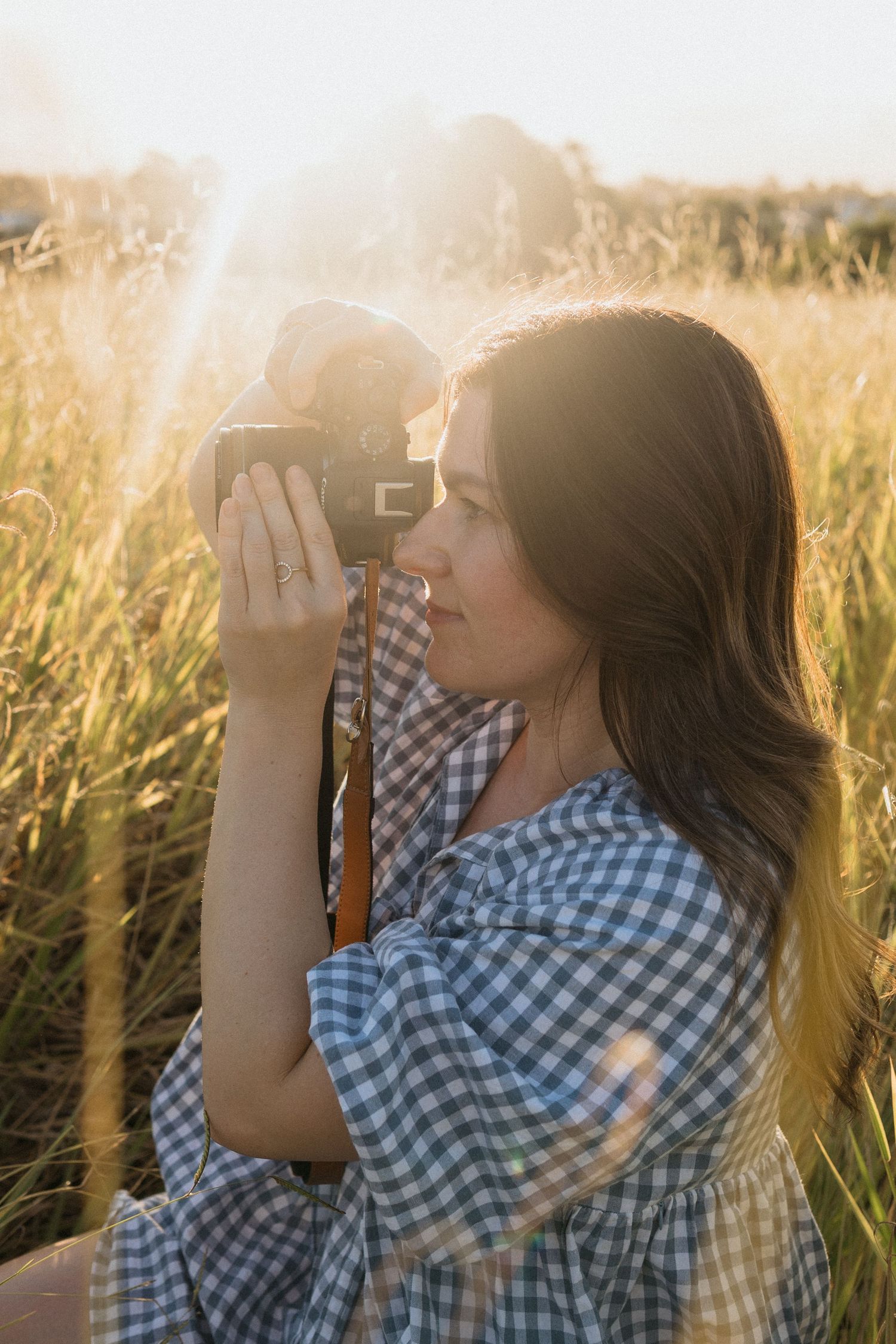 Woman in a blue gingham dress kneeling in a field and capturing photos with a camera.