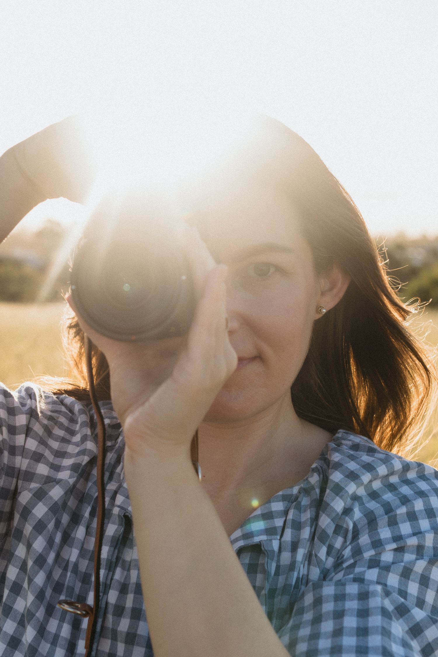 Woman in a blue gingham dress walking through a field and capturing photos with a camera.