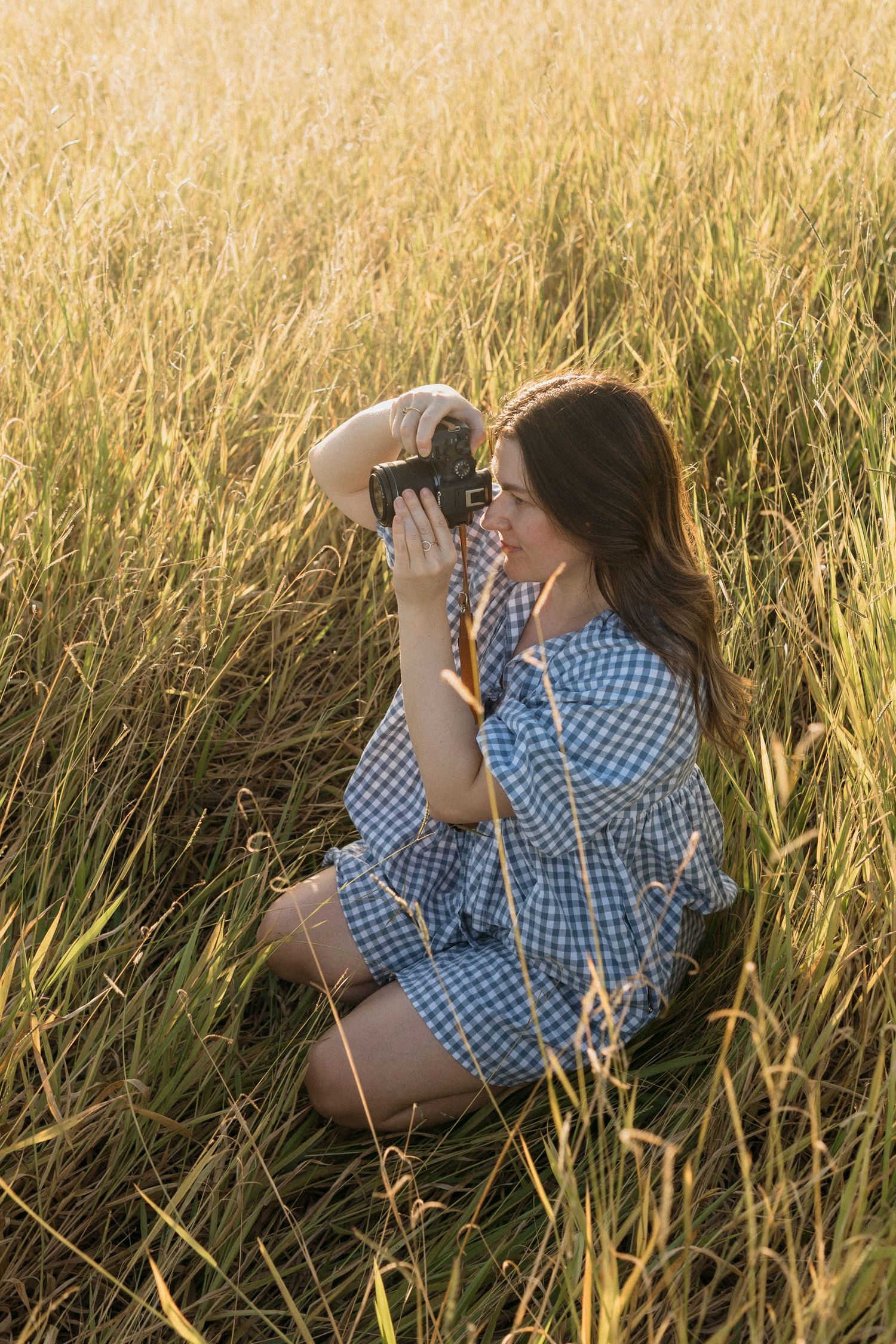 Woman in a blue gingham dress kneeling in a field and capturing photos with a camera.