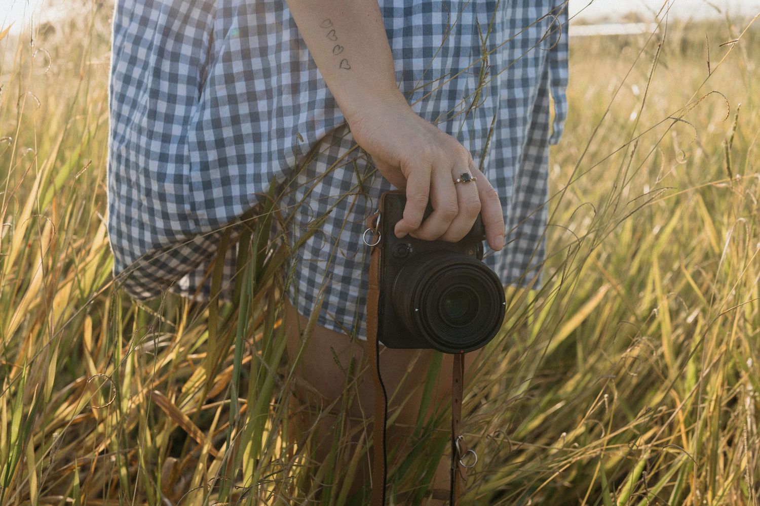 Woman in a blue gingham dress walking through a field and capturing photos with a camera.