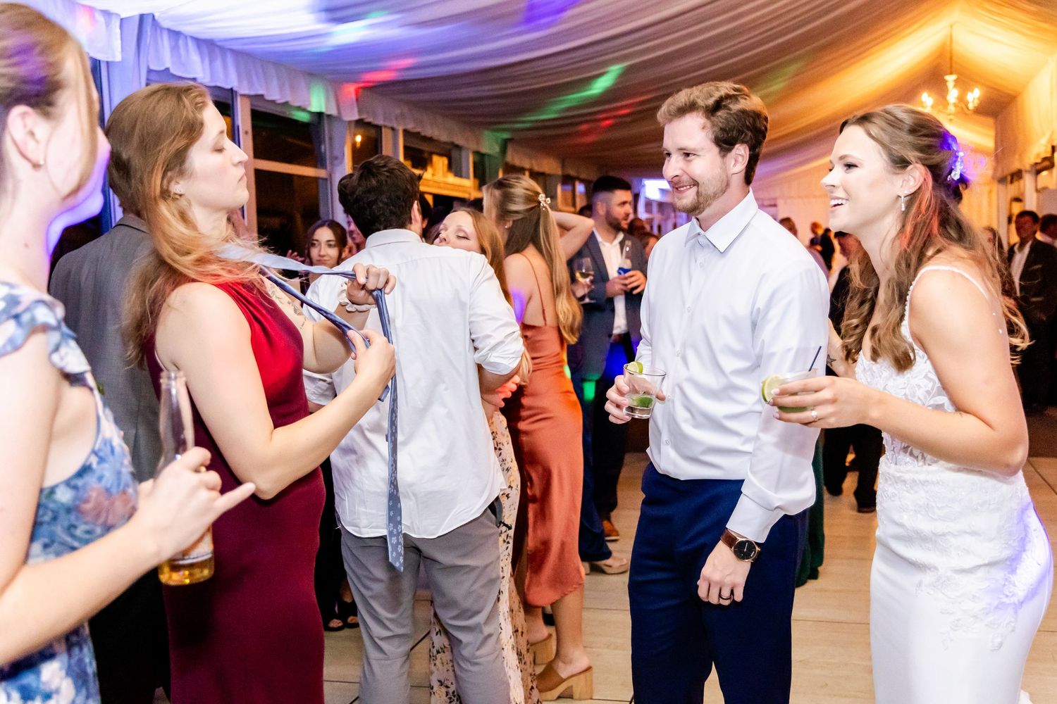 People dancing and socializing at a lively wedding reception under a tent with colorful lighting.