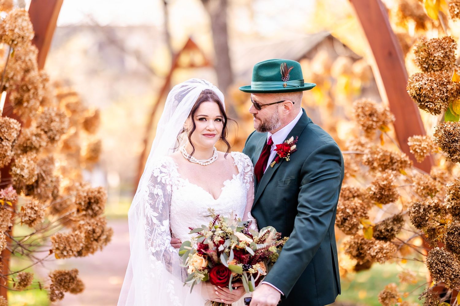 A wedding couple poses outdoors with fall foliage and warm lighting in the background.