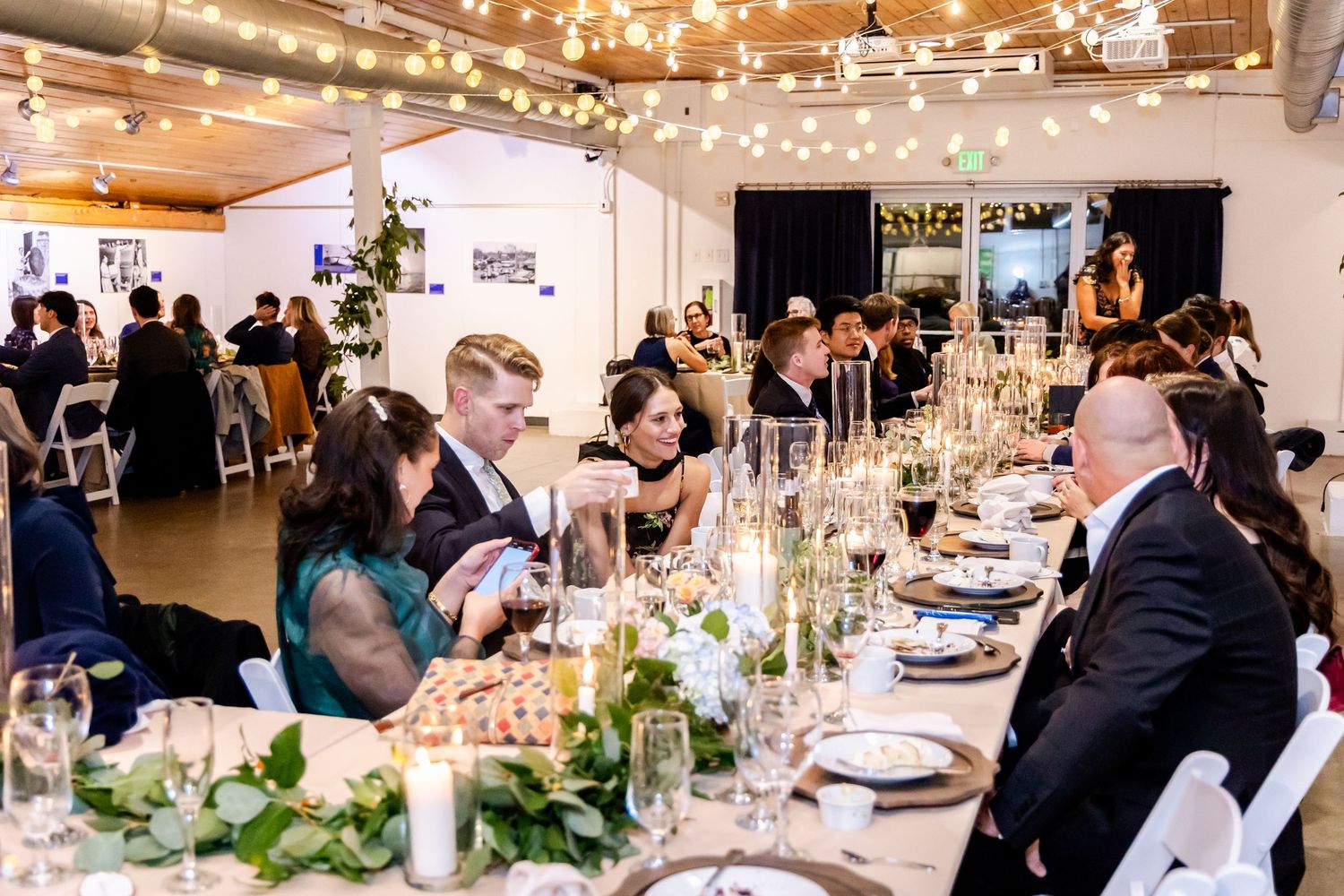 Elegant wedding reception with guests dining at long tables adorned with greenery centerpieces under string lights.