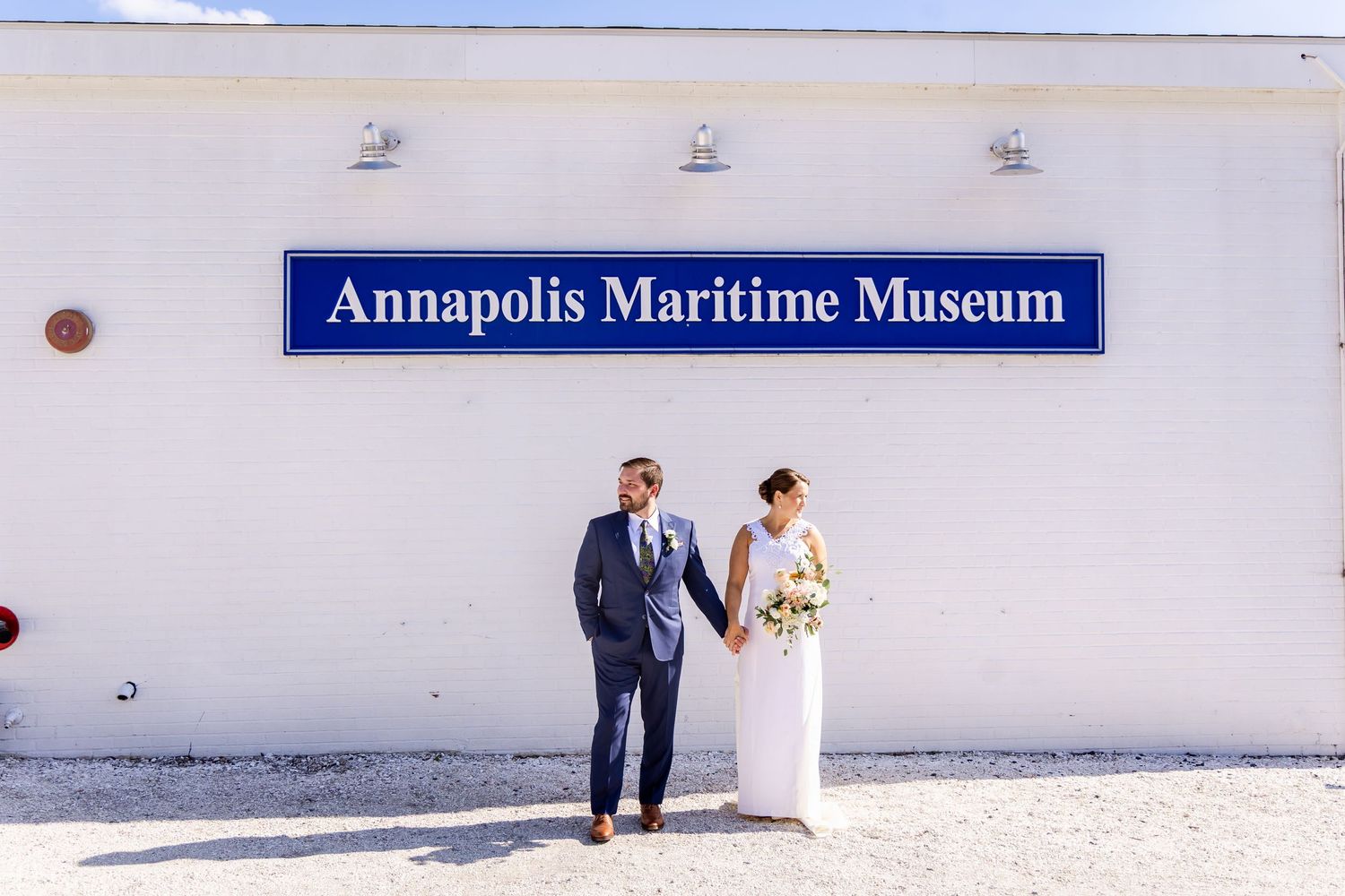 Newlyweds hold hands in front of the Annapolis Maritime Museum, bride in white gown, groom in blue suit.