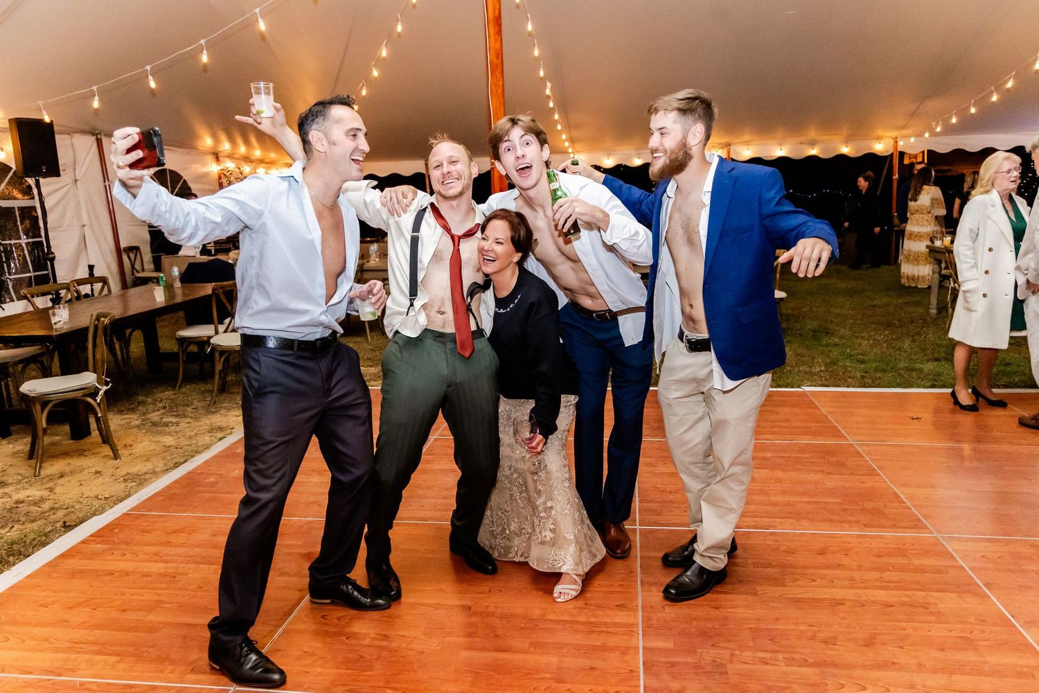 Wedding guests dance and celebrate on a wooden dance floor under a glowing tent at an evening reception.