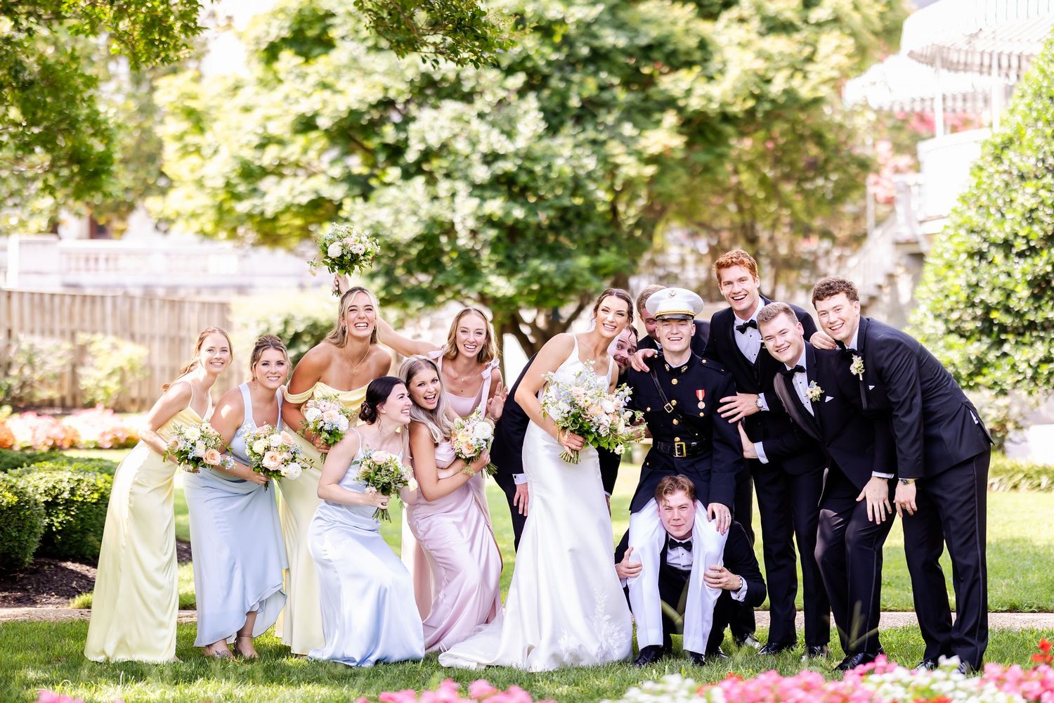 Wedding party members pose together outdoors in formal attire with bridesmaids in pink dresses and groomsmen in dark suits.