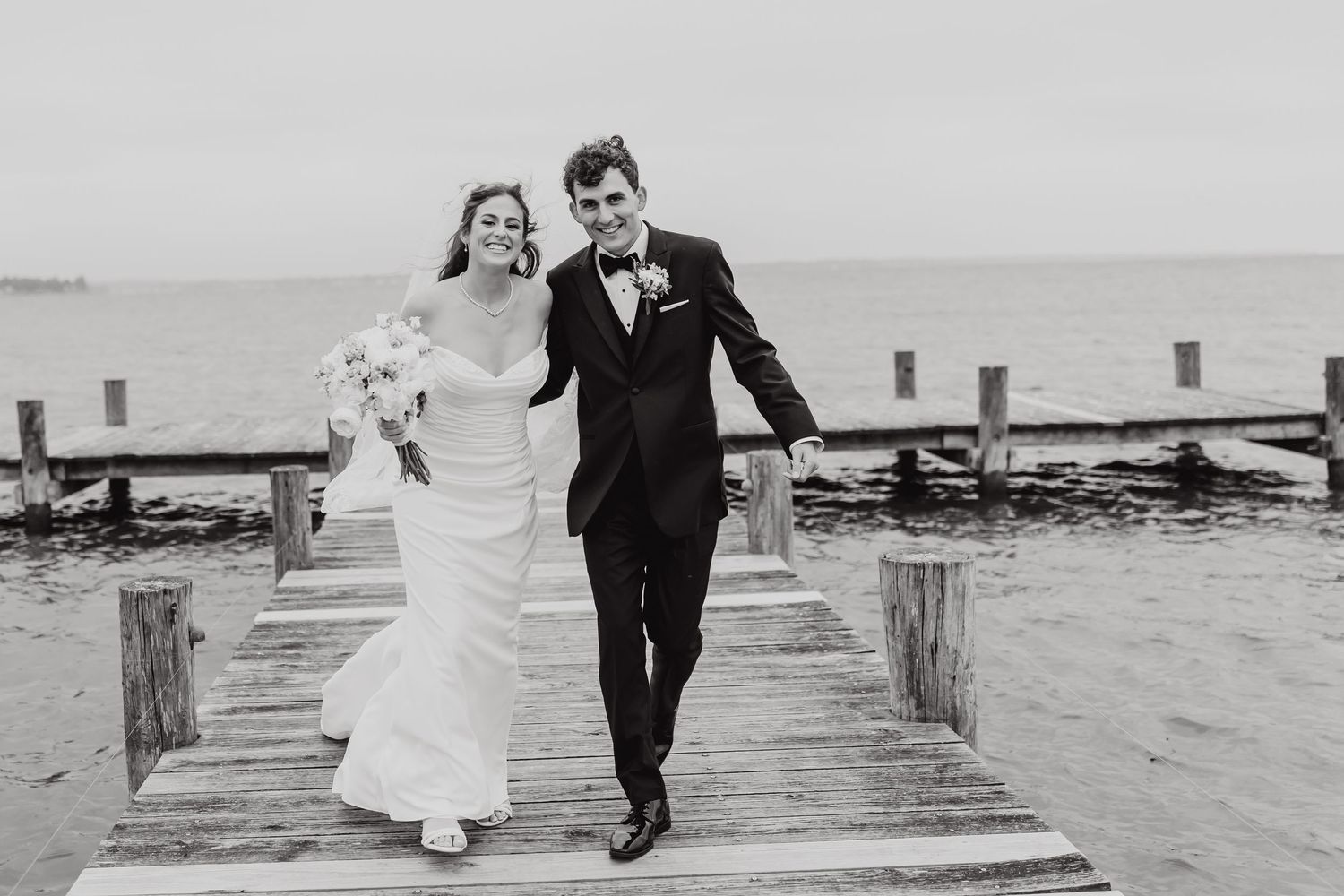 A couple in wedding attire walks together on a wooden pier over water in a series of black and white photos.