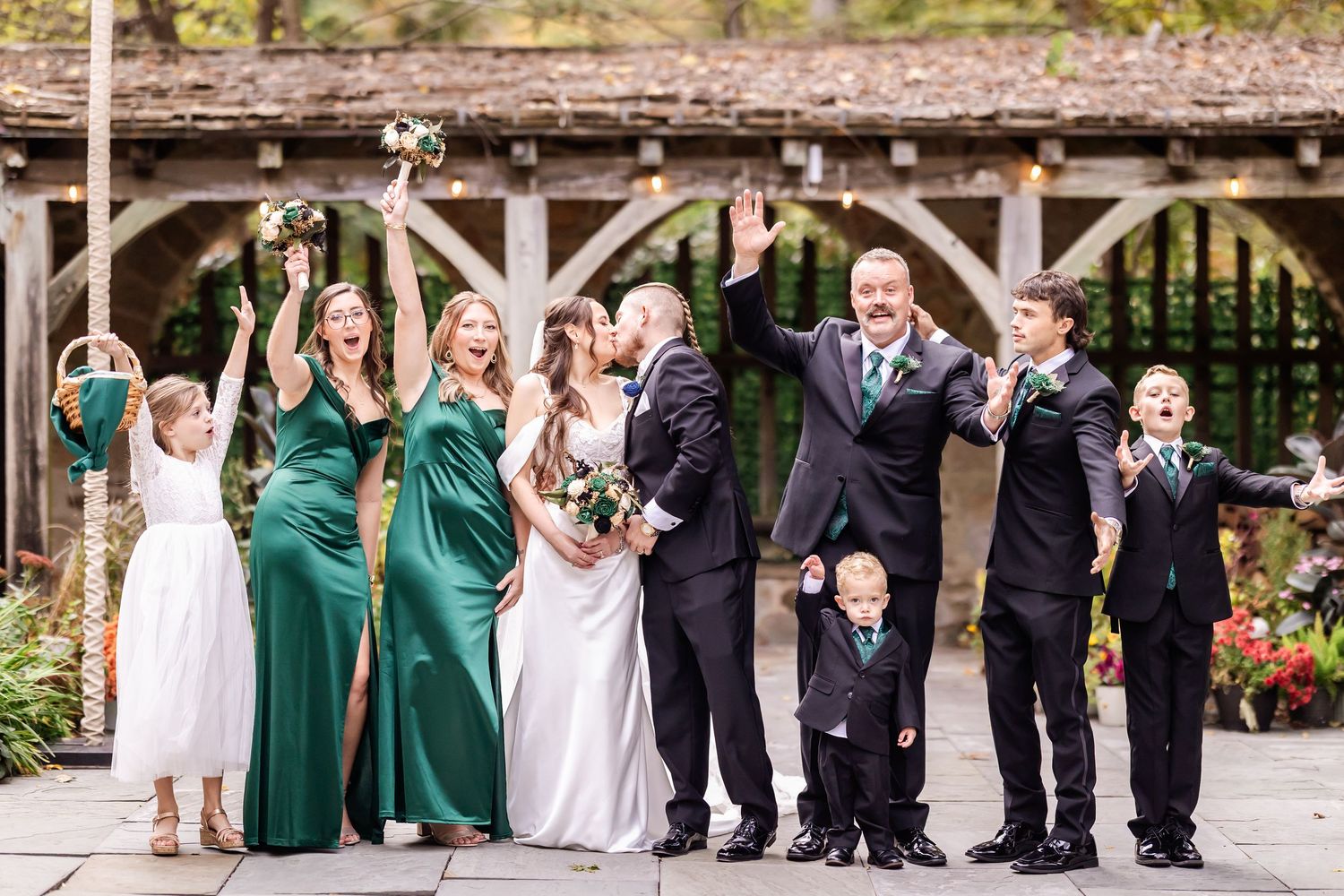 A wedding party in formal attire poses together on a patio with black and emerald green dresses and suits.