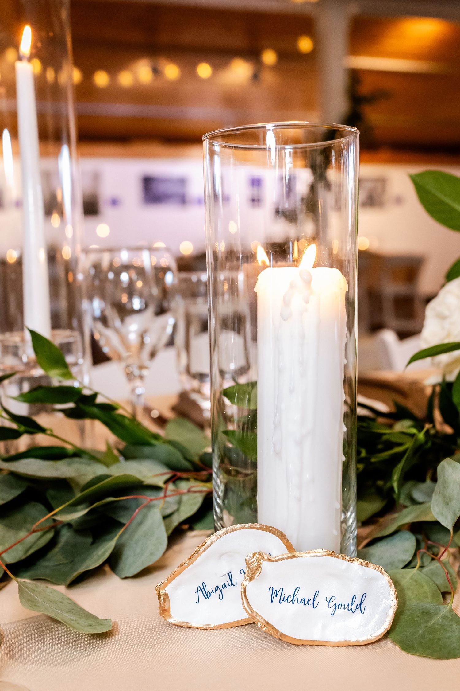 Elegant wedding table setting with white candle in glass cylinder, eucalyptus leaves, and wood slice name cards.