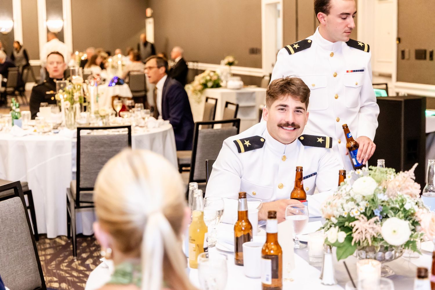 Wedding reception guests enjoying dinner at elegantly decorated tables with floral centerpieces and white linens.