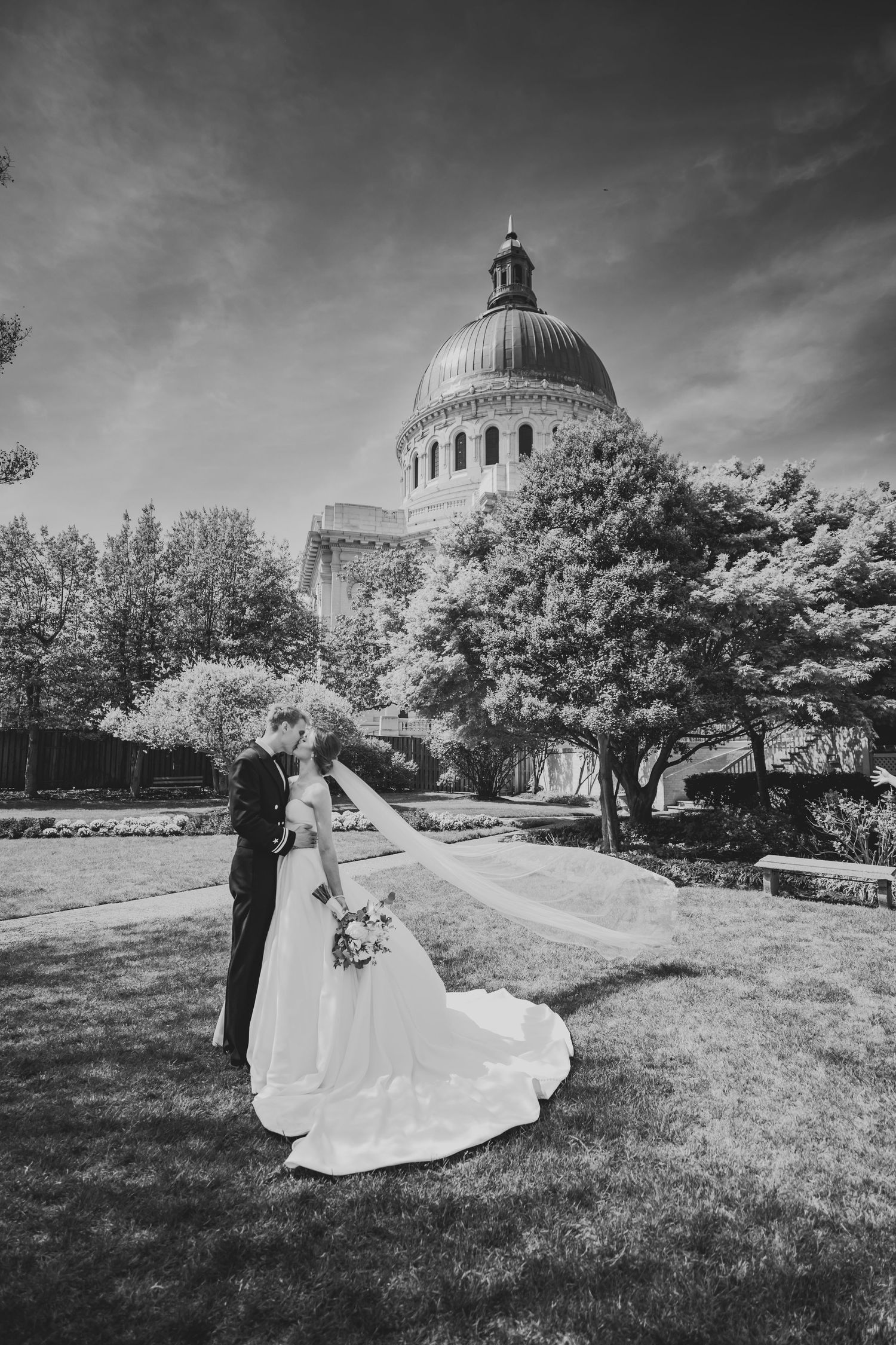 A wedding couple stands on a grassy lawn with a domed building in the background under dramatic cloudy skies.