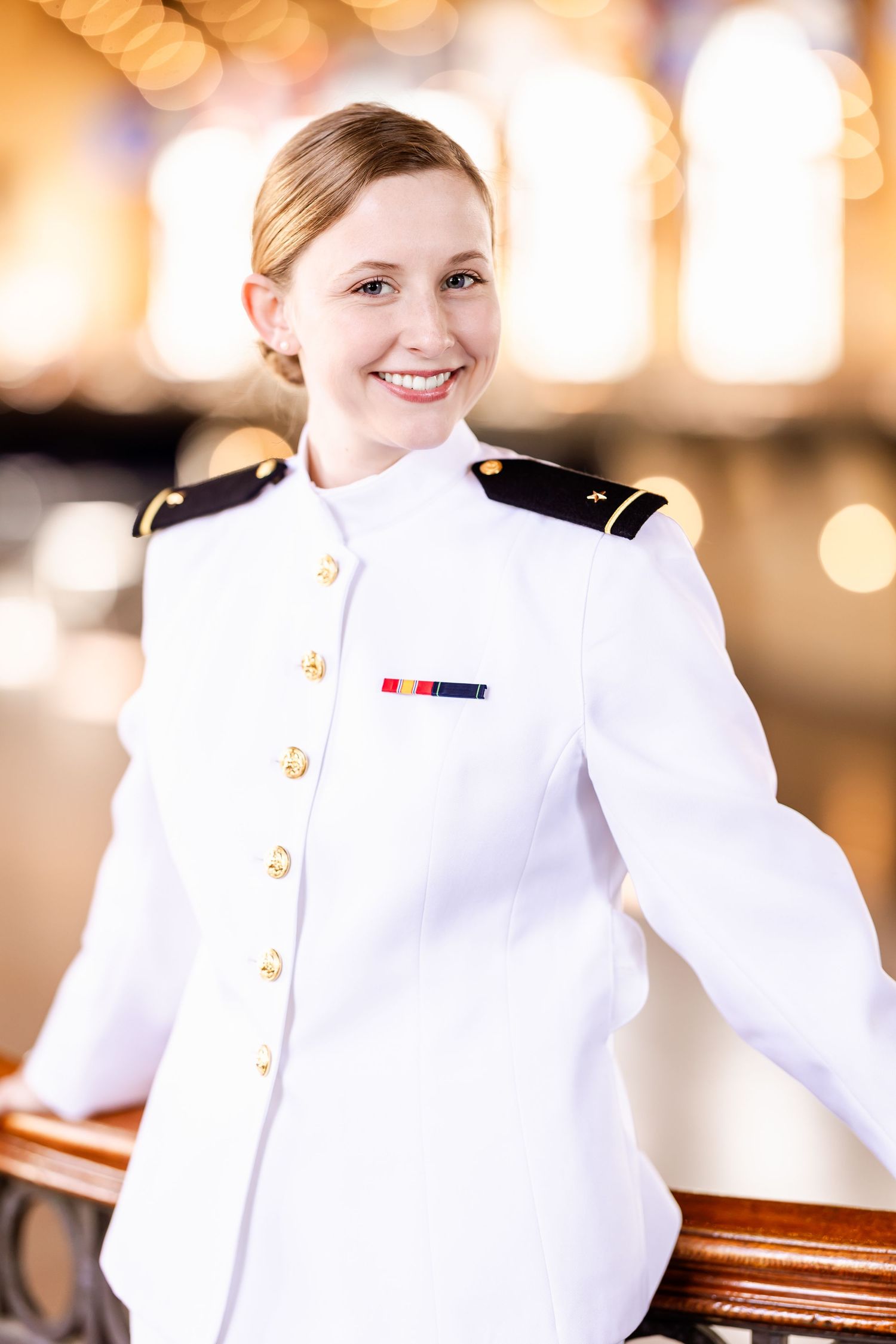 Naval officer in dress white uniform smiling while standing in an elegant venue with soft bokeh lighting.
