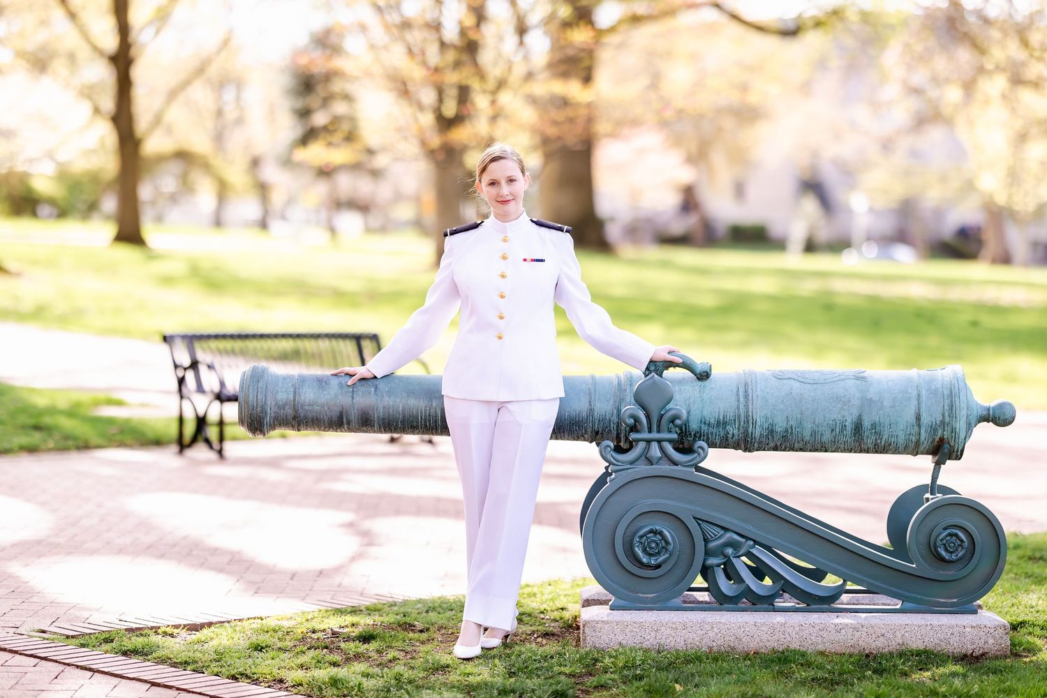 A naval officer in white dress uniform poses beside a historic cannon display in a sunny park setting.