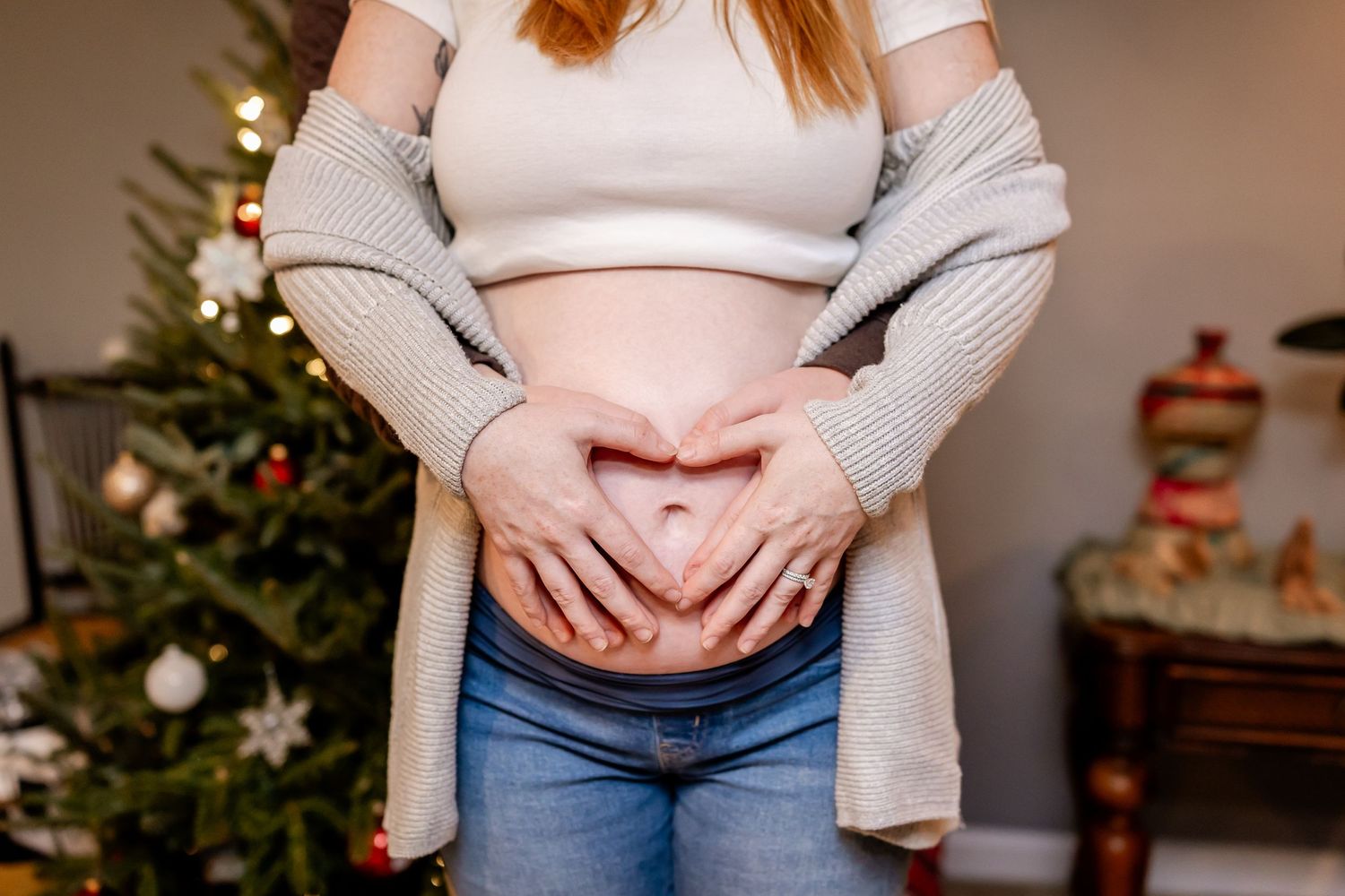 A casual holiday scene showing a person in a gray cardigan and white top making a heart shape with hands over their belly.
