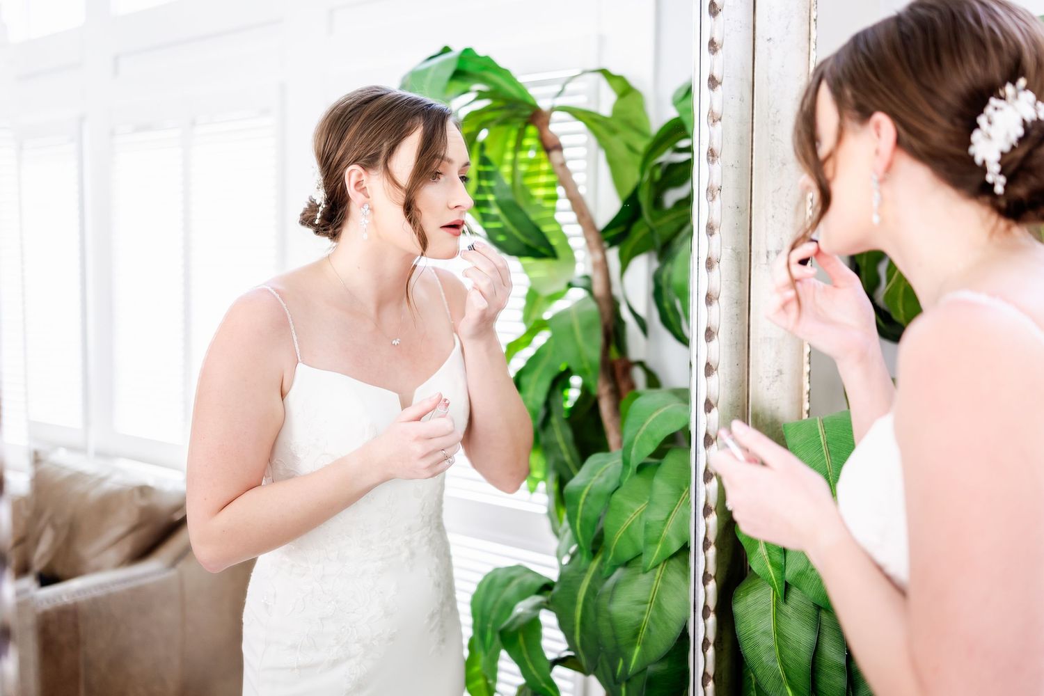 A person in a white wedding dress looks at their reflection in a mirror with tropical plants in the background.