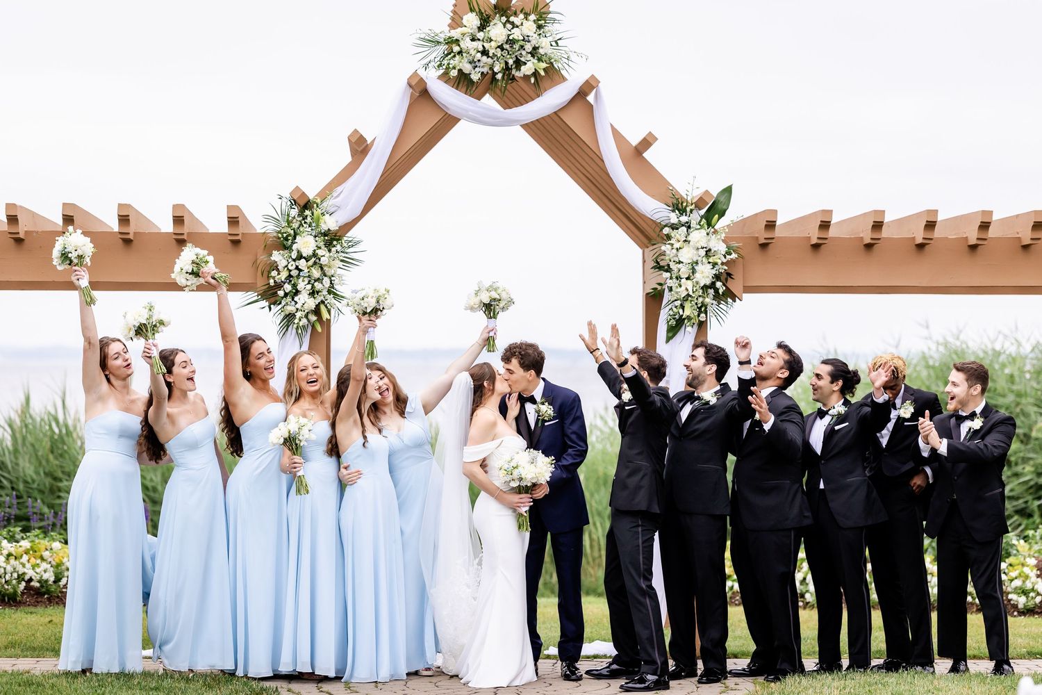 Wedding party celebrates under an arch with bridesmaids in blue dresses and groomsmen in black suits.