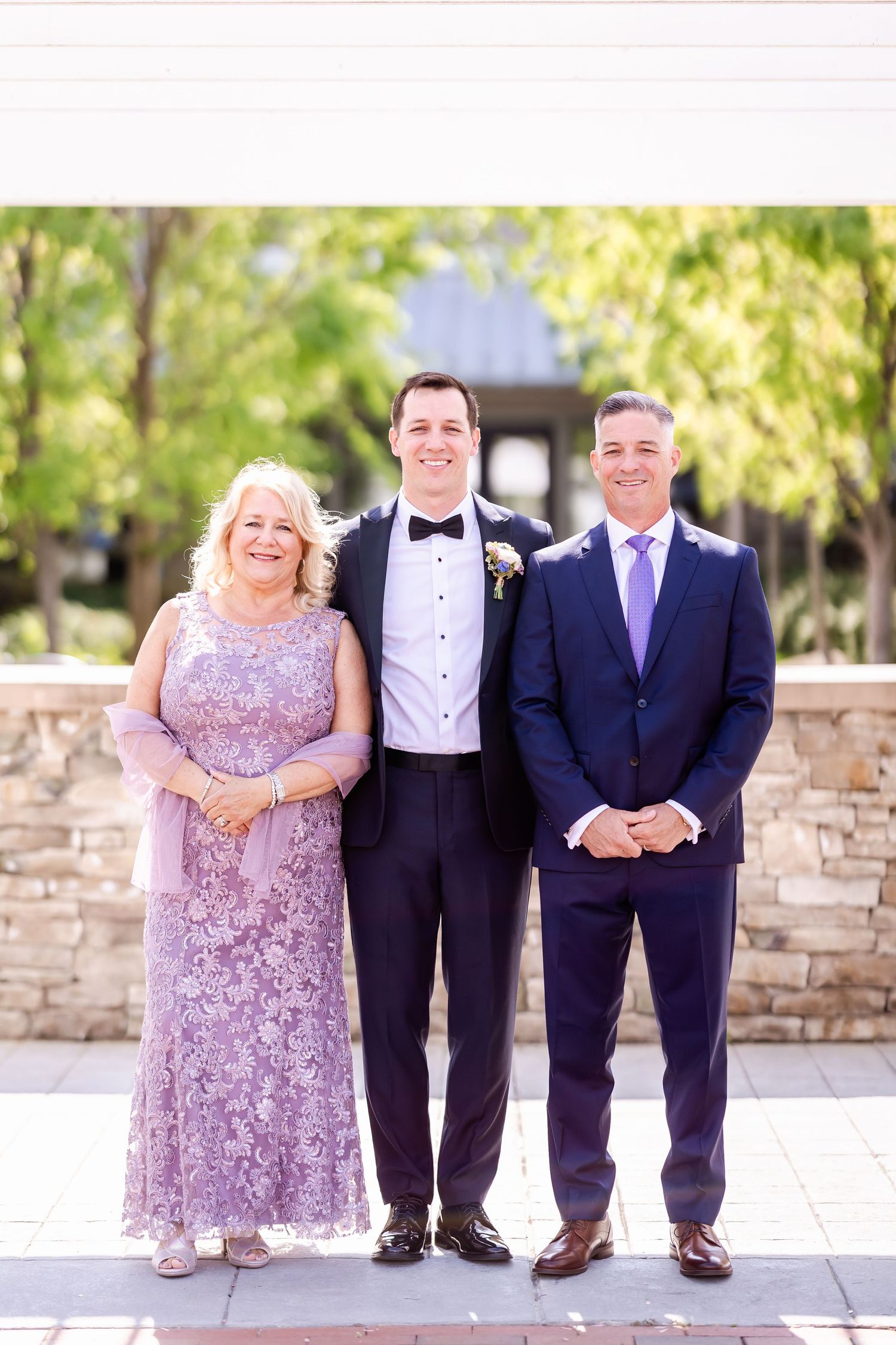 Wedding party members pose together in formal attire against a light stone wall.
