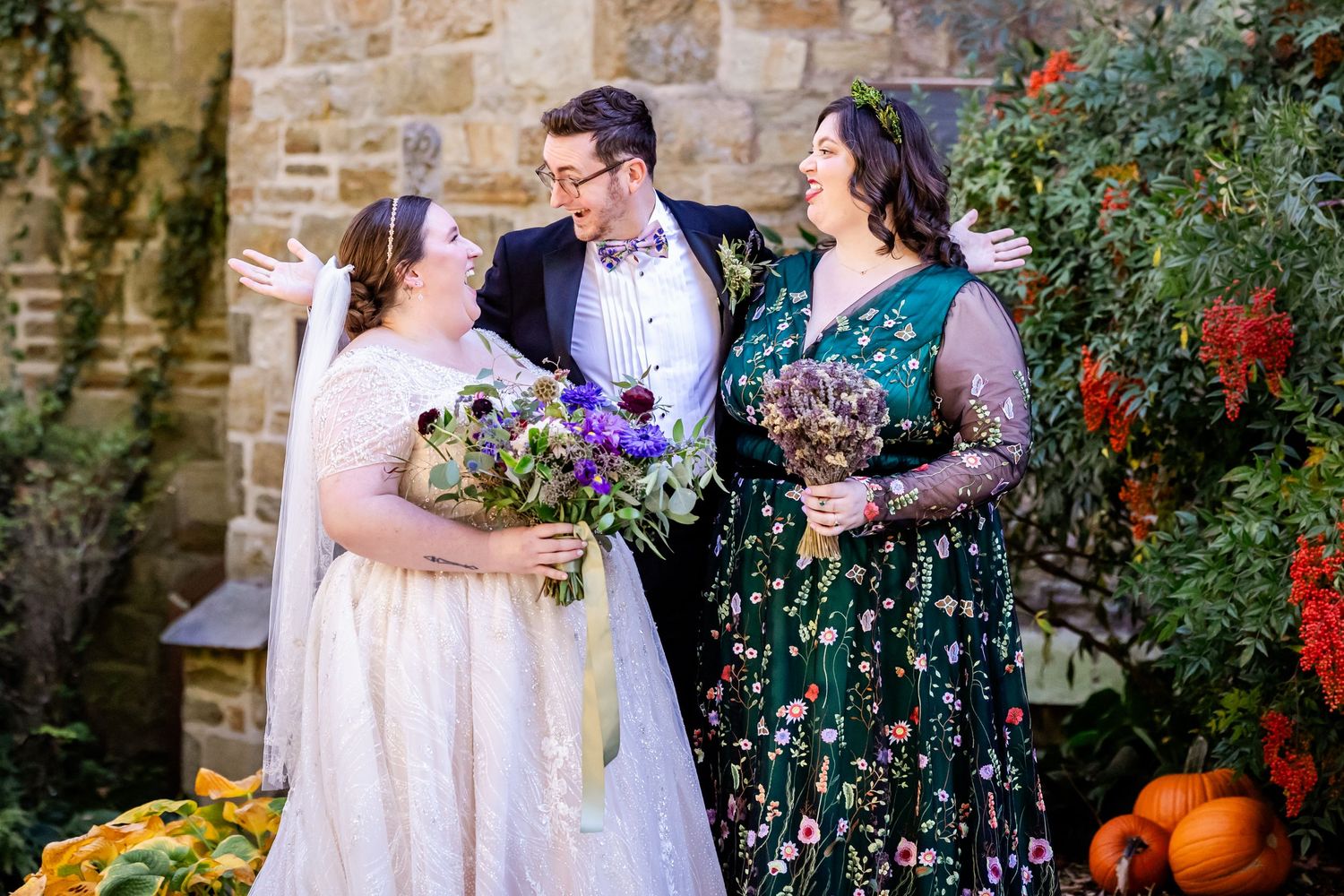 Wedding party members share laughs while holding vibrant purple bouquets against stone wall with pumpkins.