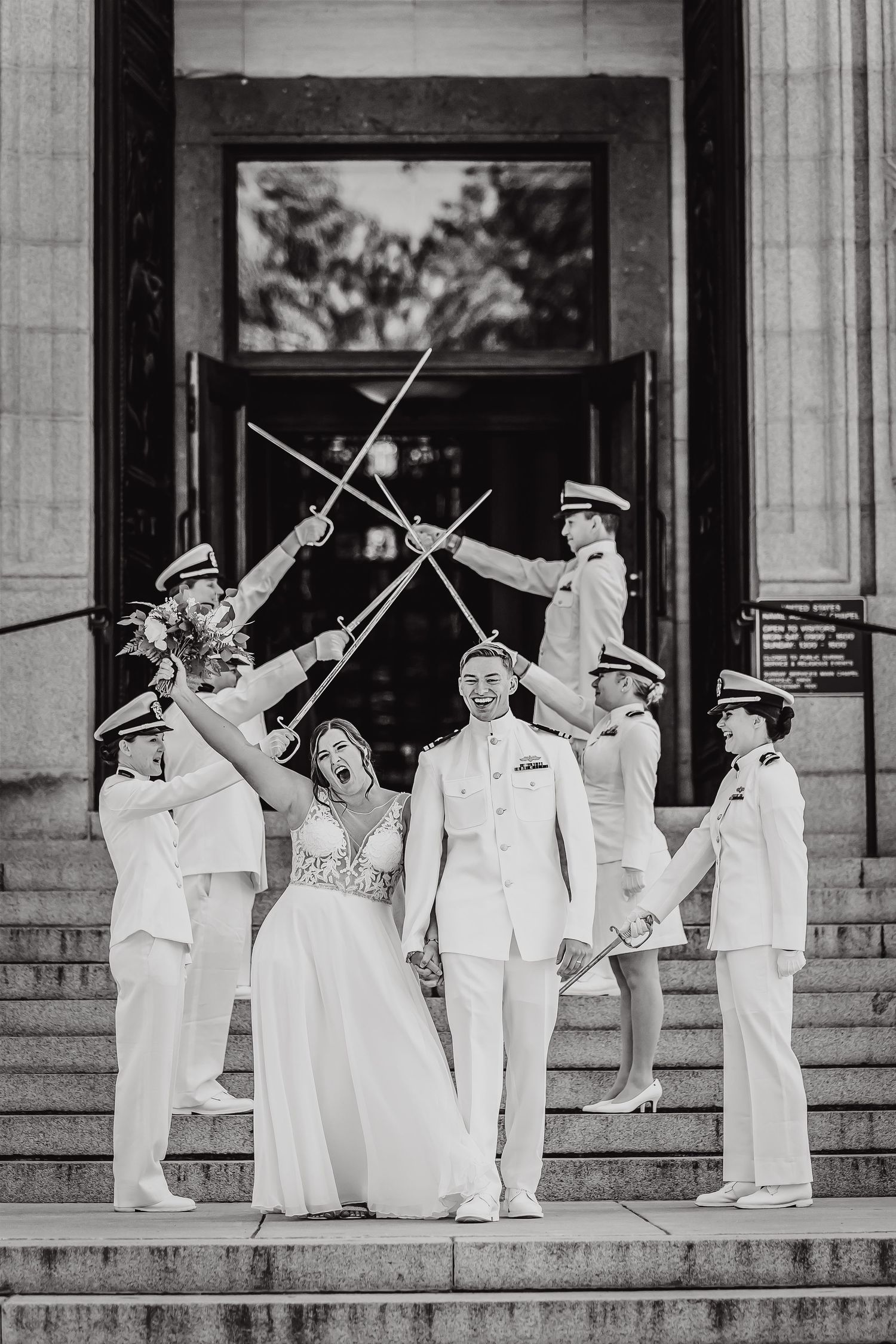 A bride and groom stand on stairs with military officers forming a traditional sword arch in black and white photography.