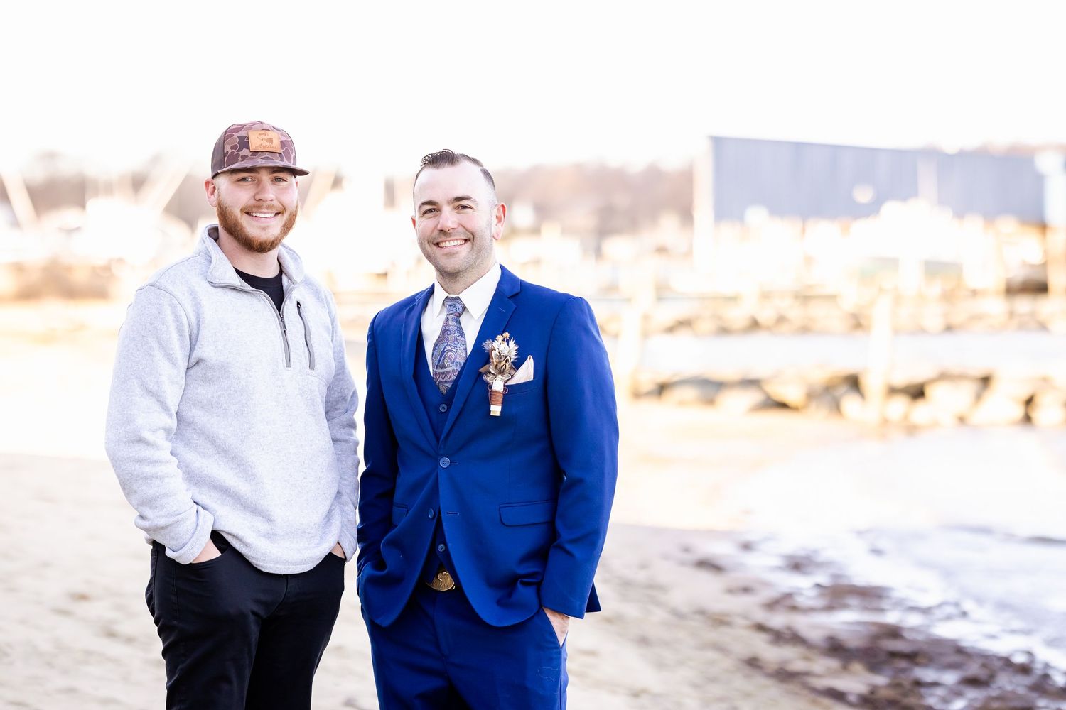 Two people stand together on a beach, one in casual wear and one in a royal blue suit with boutonniere.