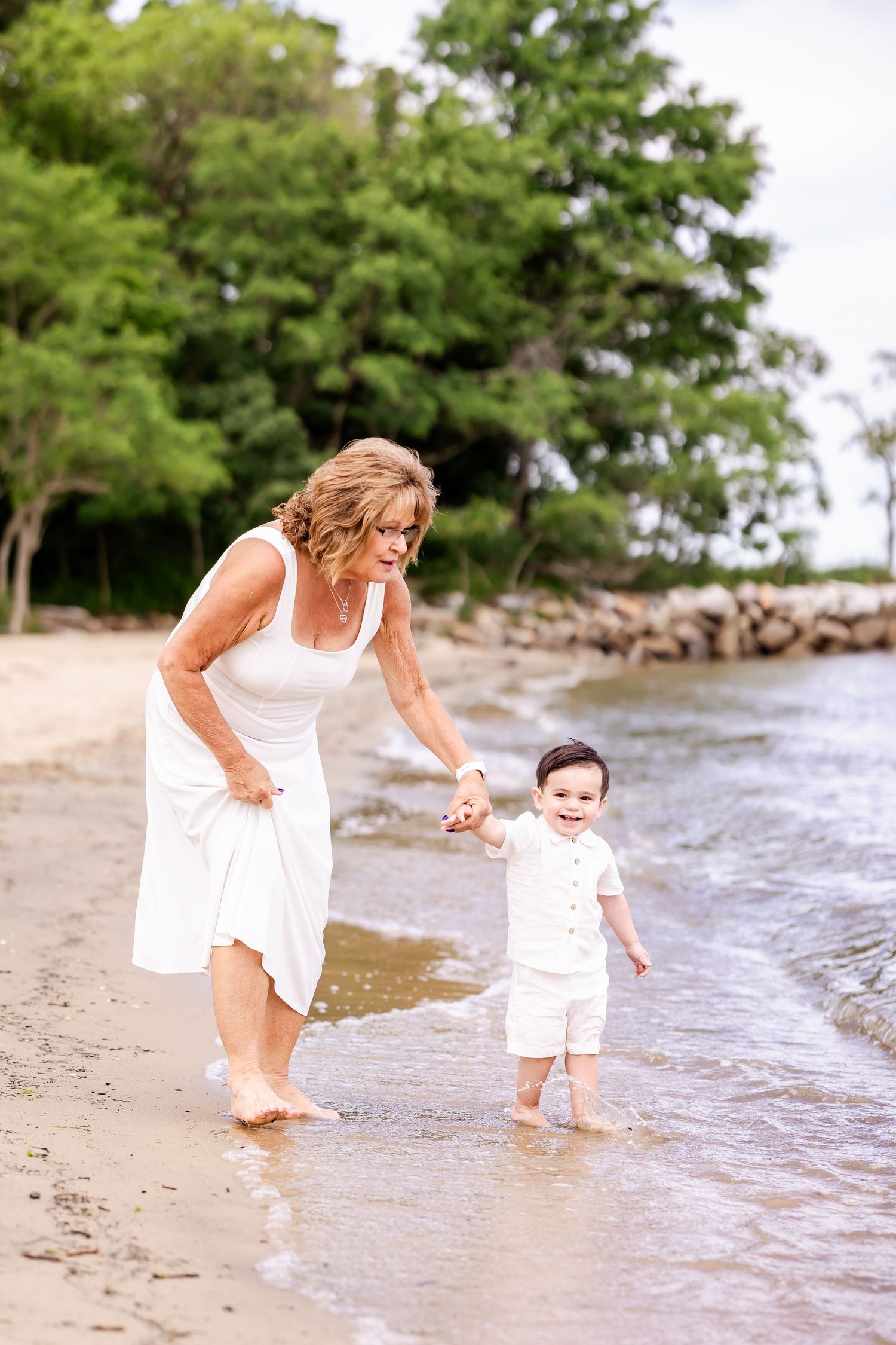 Parent and small child in white outfits splash together at the water's edge on a beach with lush trees behind them.