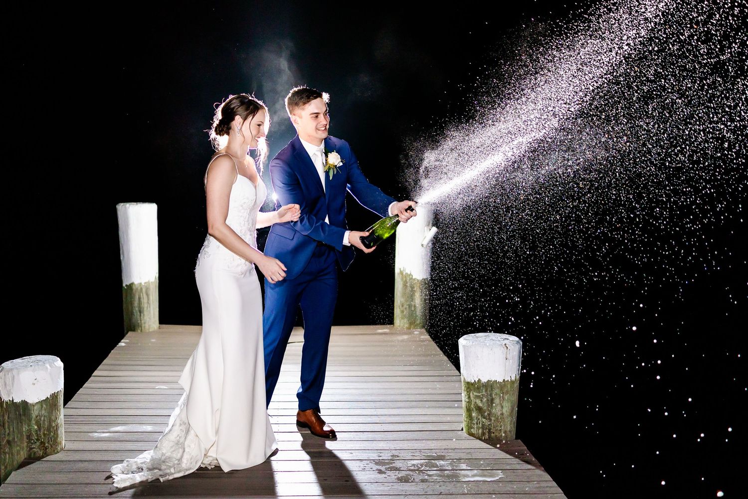 Couple sprays champagne on dock at night creating sparkling effect against dark sky.