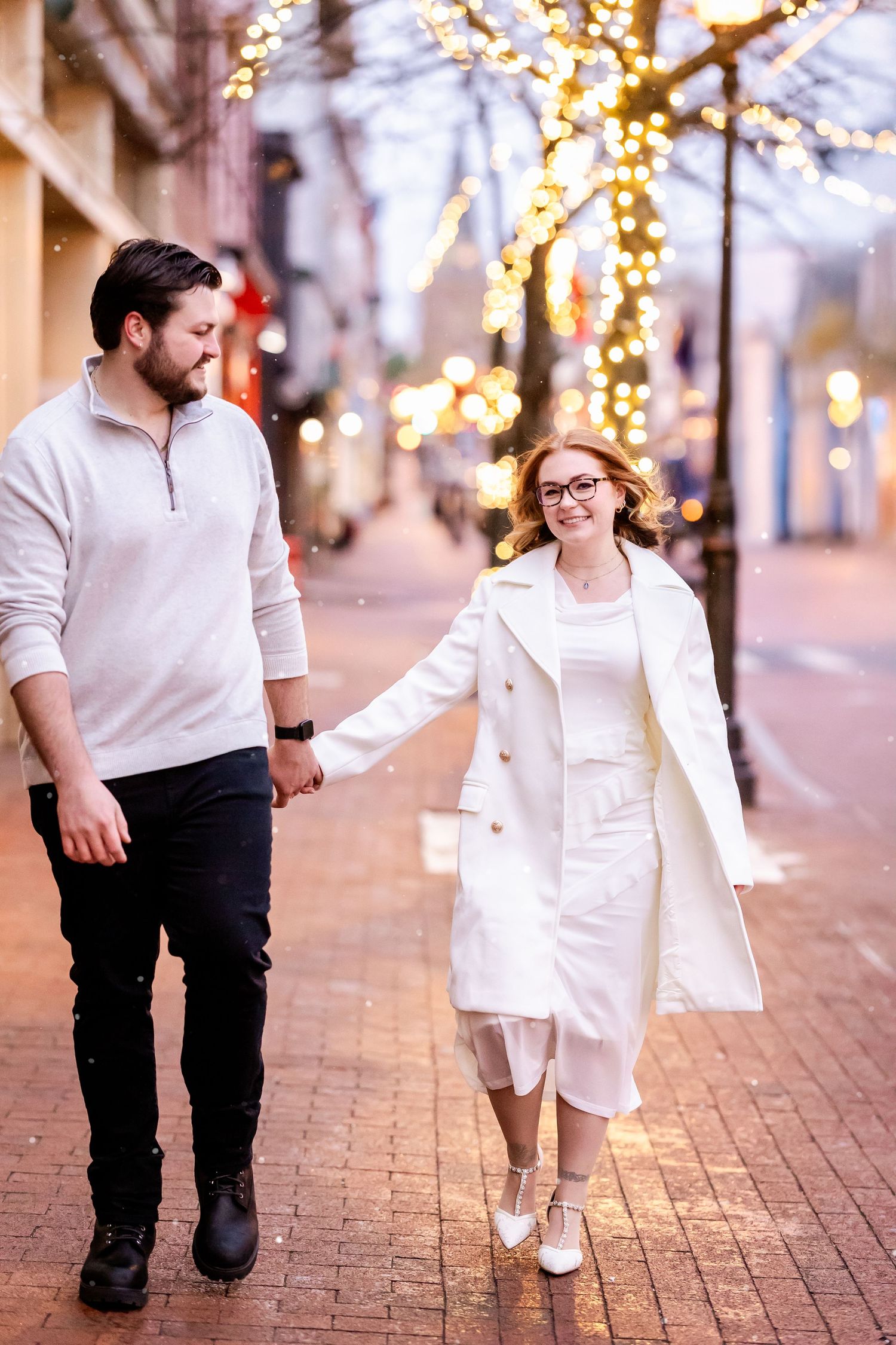 A parent and child hold hands while walking down a brick street decorated with twinkling lights at dusk.