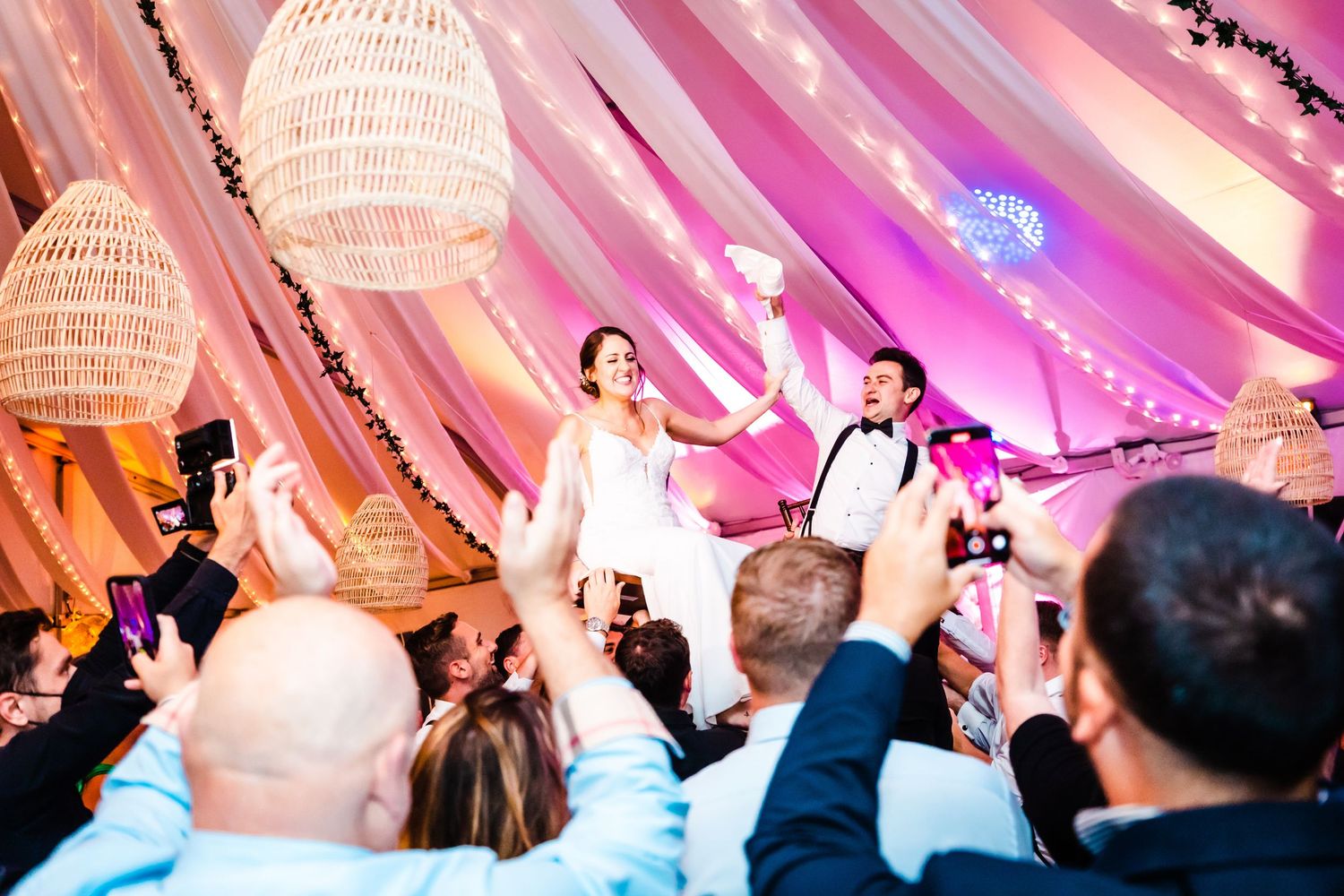 Joyful celebration at a wedding reception with guests lifting their hands under illuminated pink decor and chandeliers.