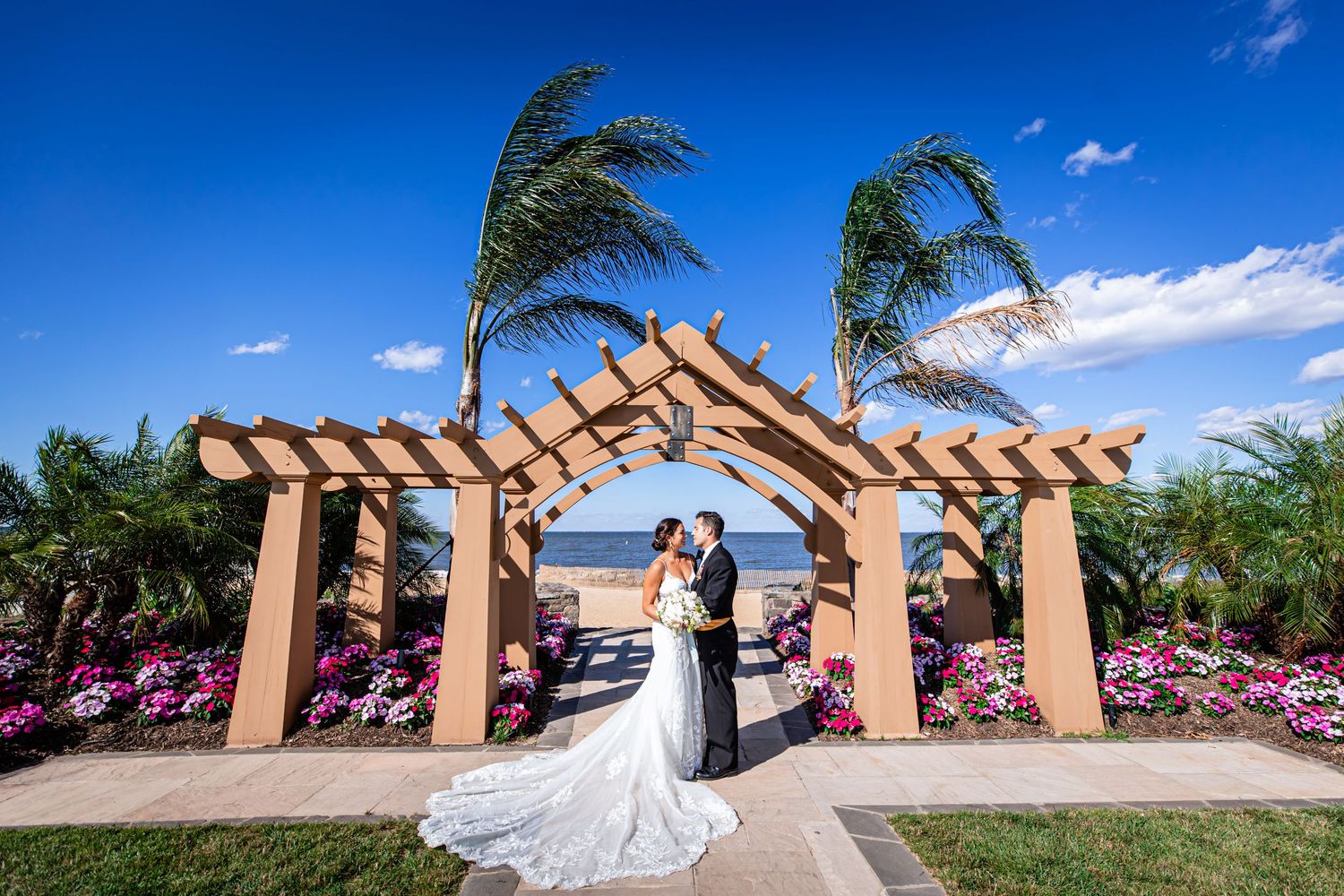 Couple shares romantic moment under wooden pergola with swaying palm trees and pink flowers by water.
