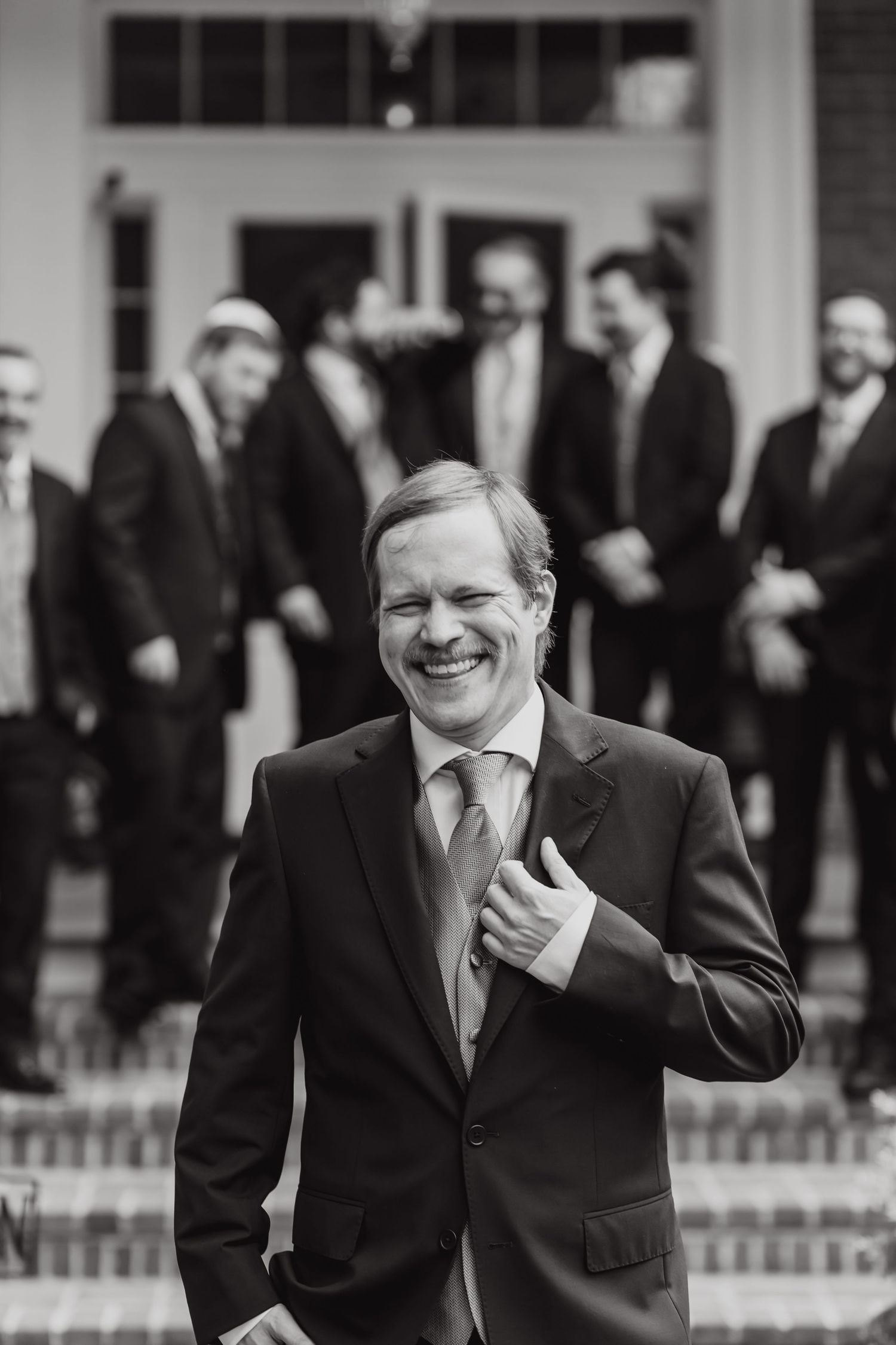 Black and white photo of person in suit smiling joyfully and making a hand gesture while standing outdoors with others behind.