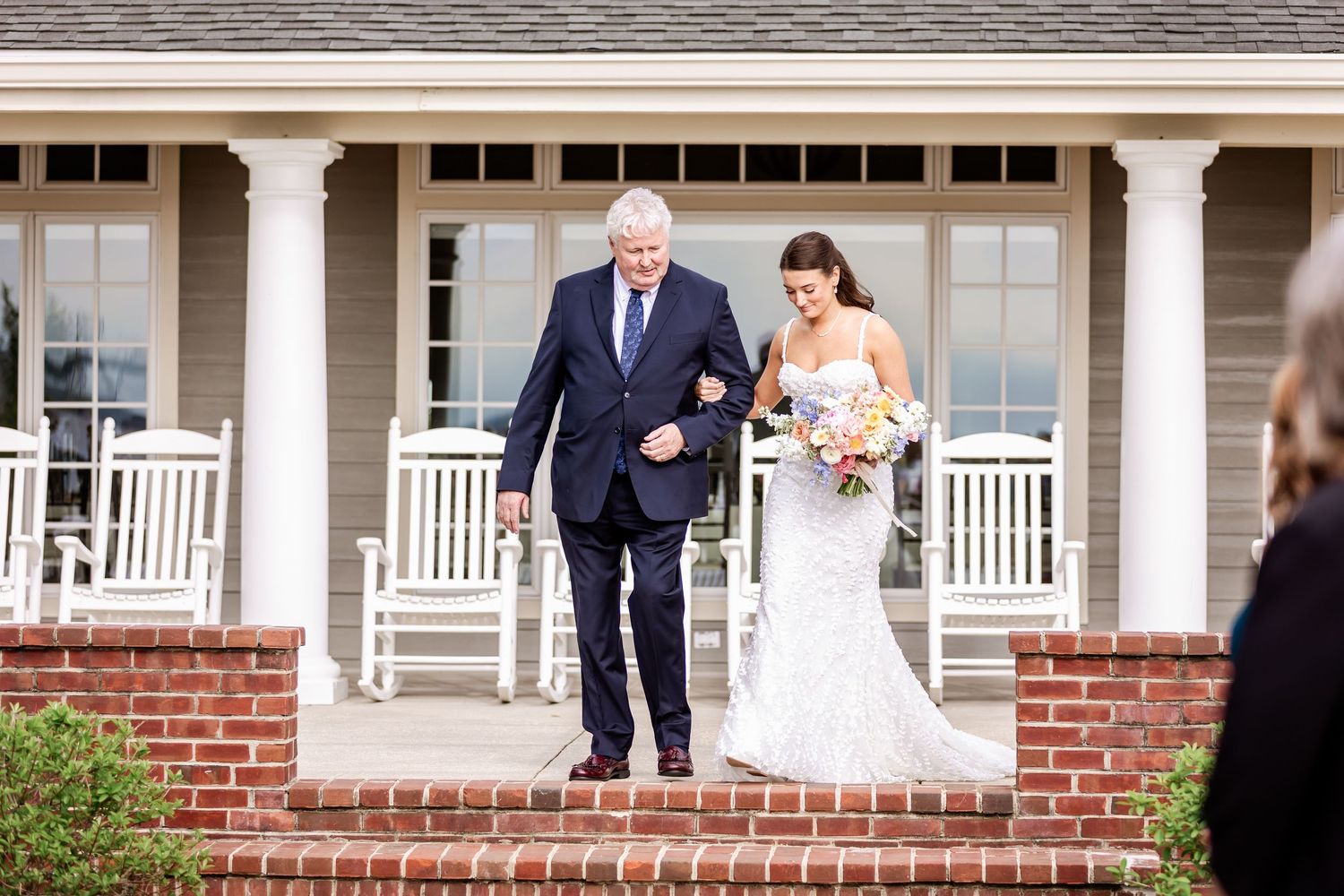 A bride in a fitted lace gown walks down a columned porch with an escort.