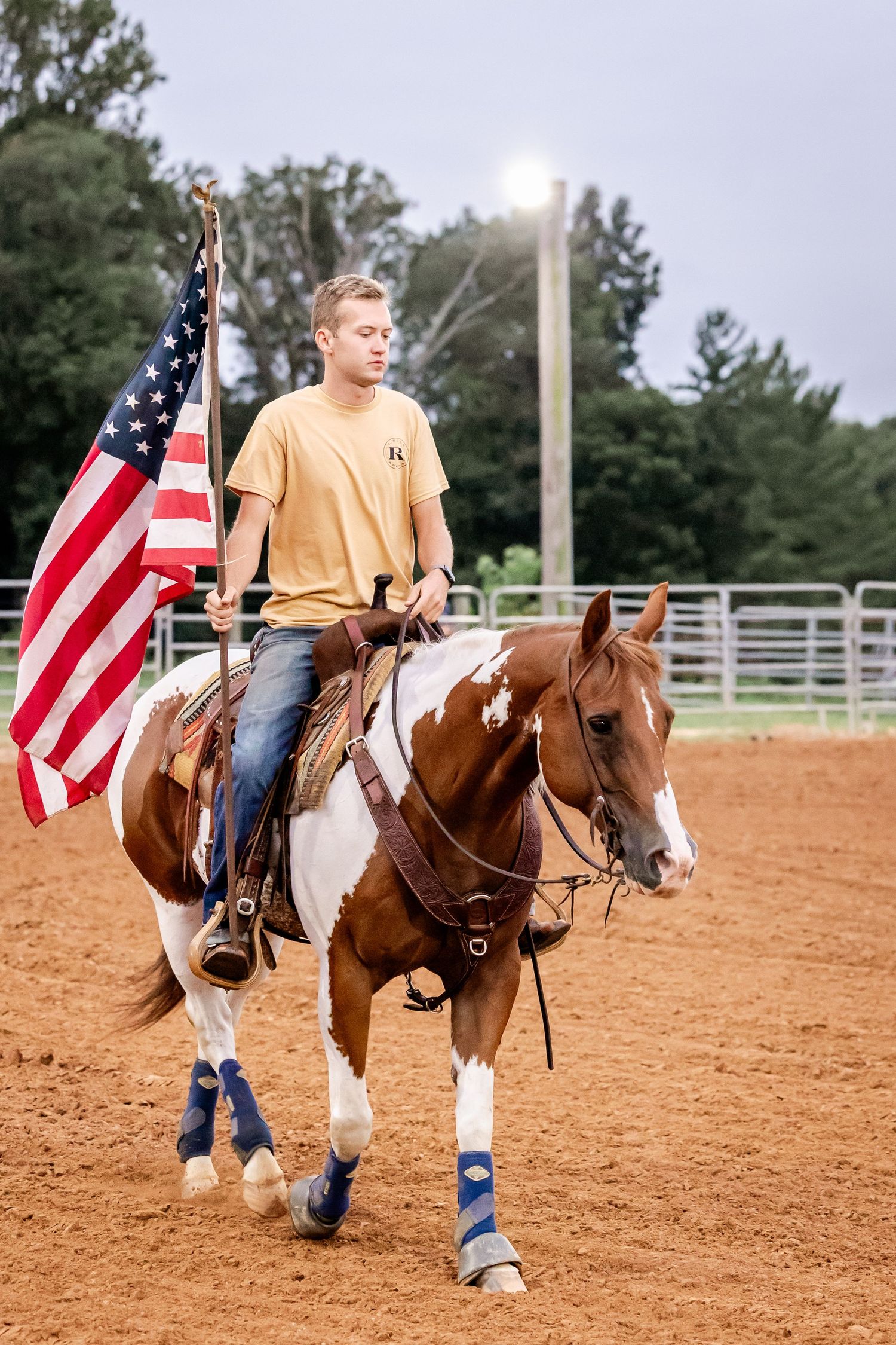 Horseback rider carrying American flag on paint horse in sandy arena.