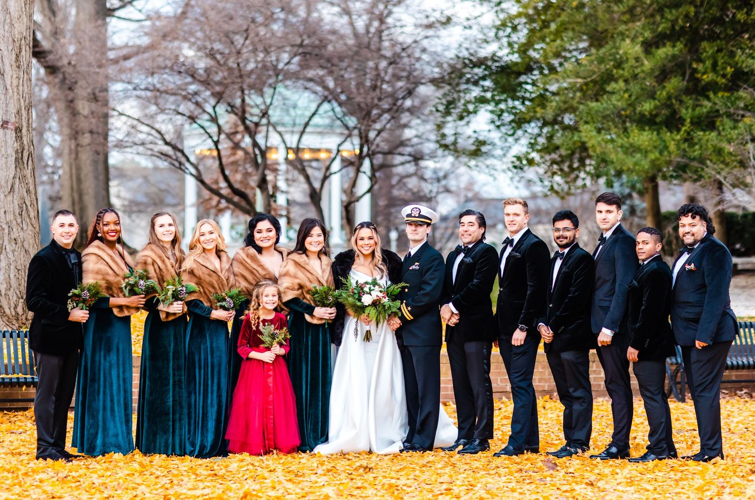 Wedding party stands in formal attire on fallen autumn leaves with bridesmaids in teal dresses and groomsmen in dark suits.