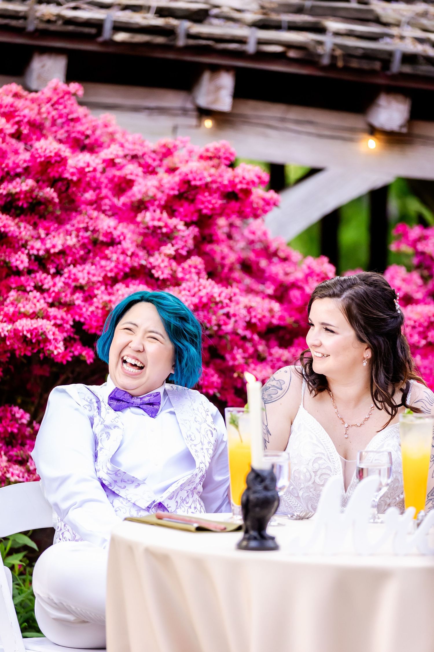 Two people share a laugh while sitting at an outdoor table with bright pink flowers in the background.