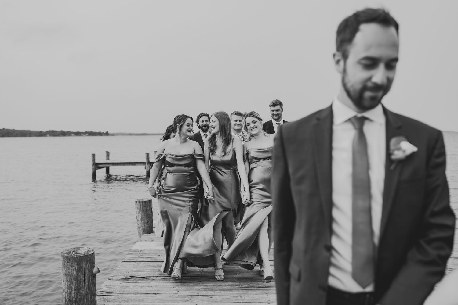 A wedding party walks along a wooden dock by a lake while getting photographed in black and white.