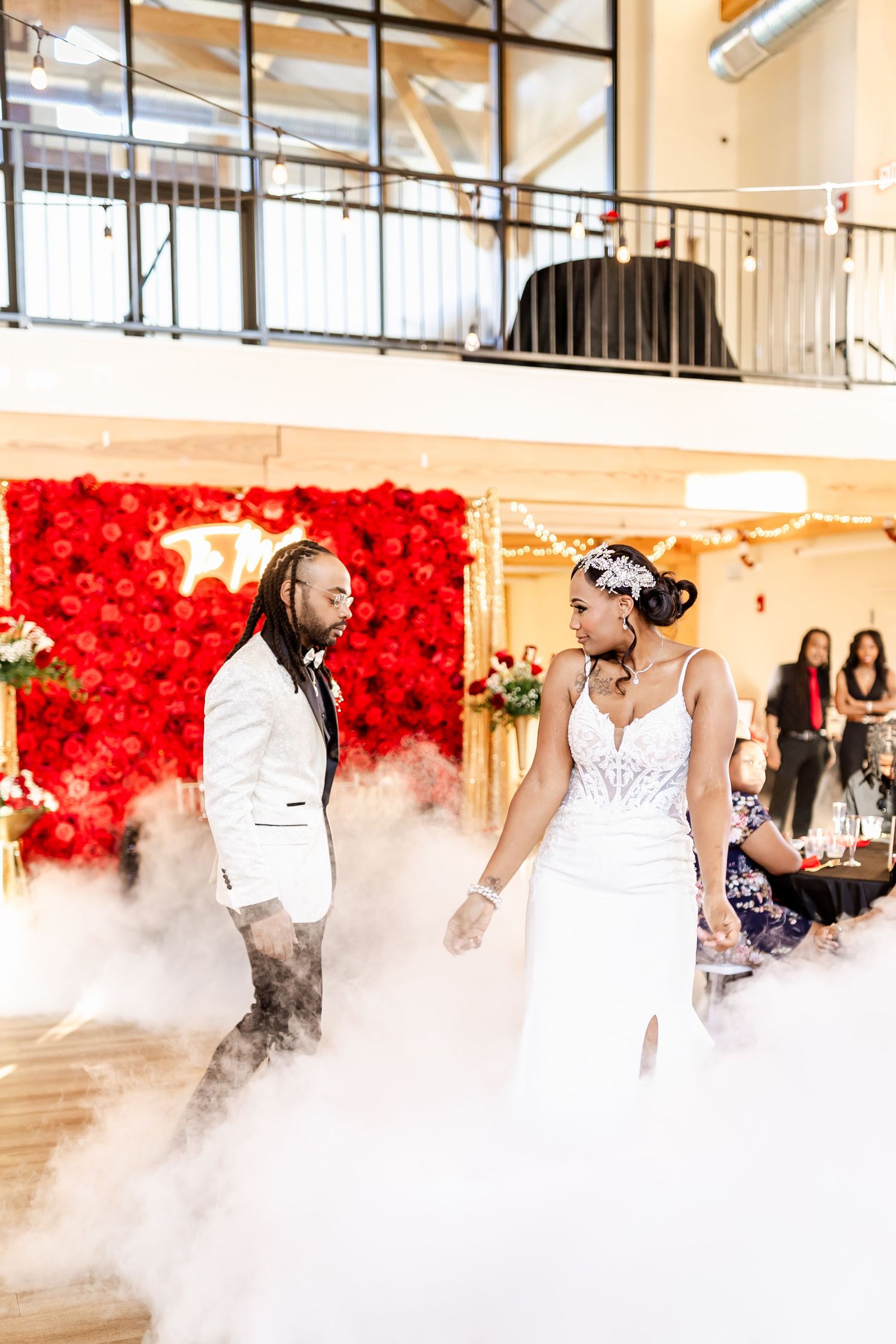 Wedding couple sharing a dance moment surrounded by atmospheric fog and bright red floral backdrop.