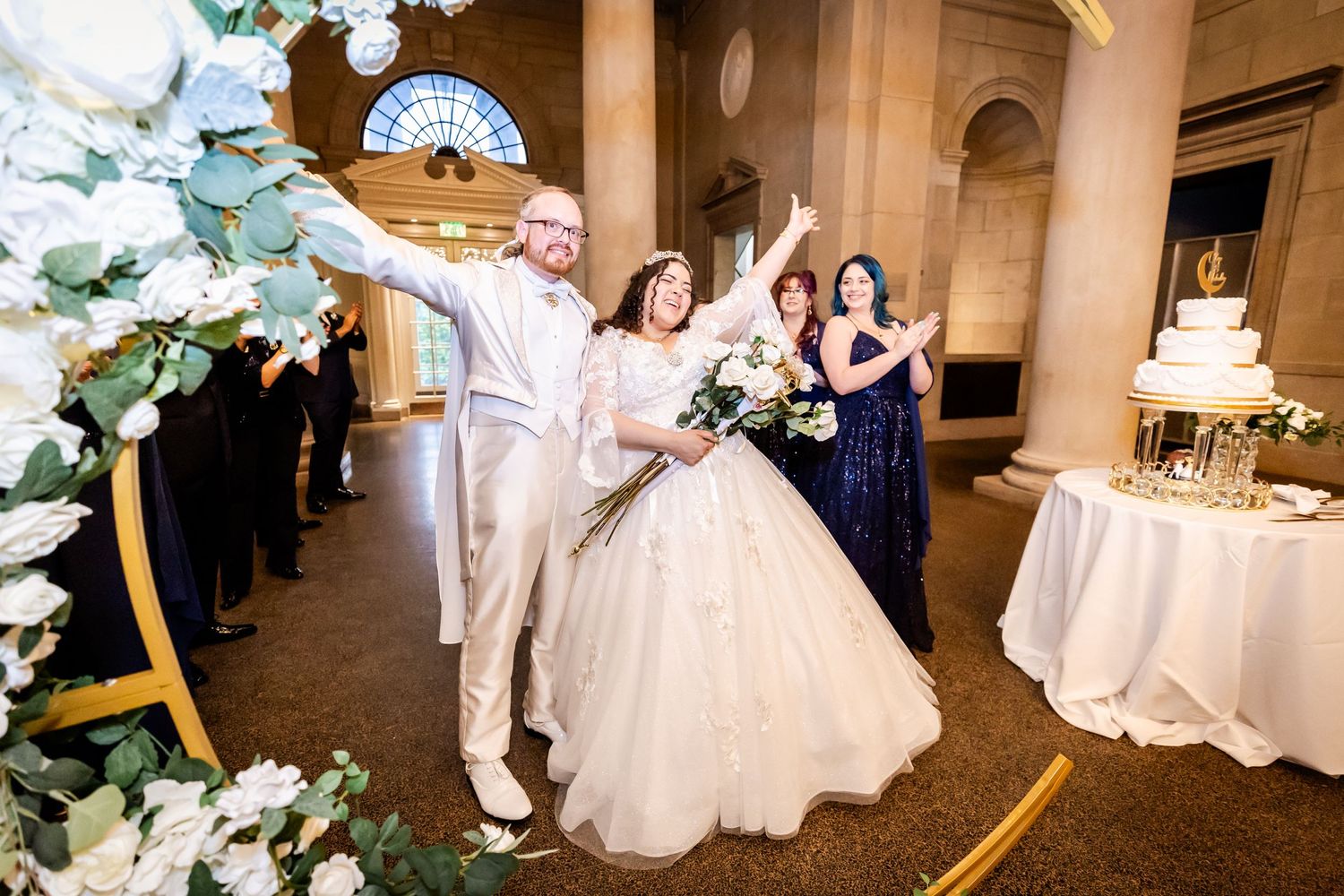 Couple celebrates with arms raised during wedding reception under floral arch and columns.