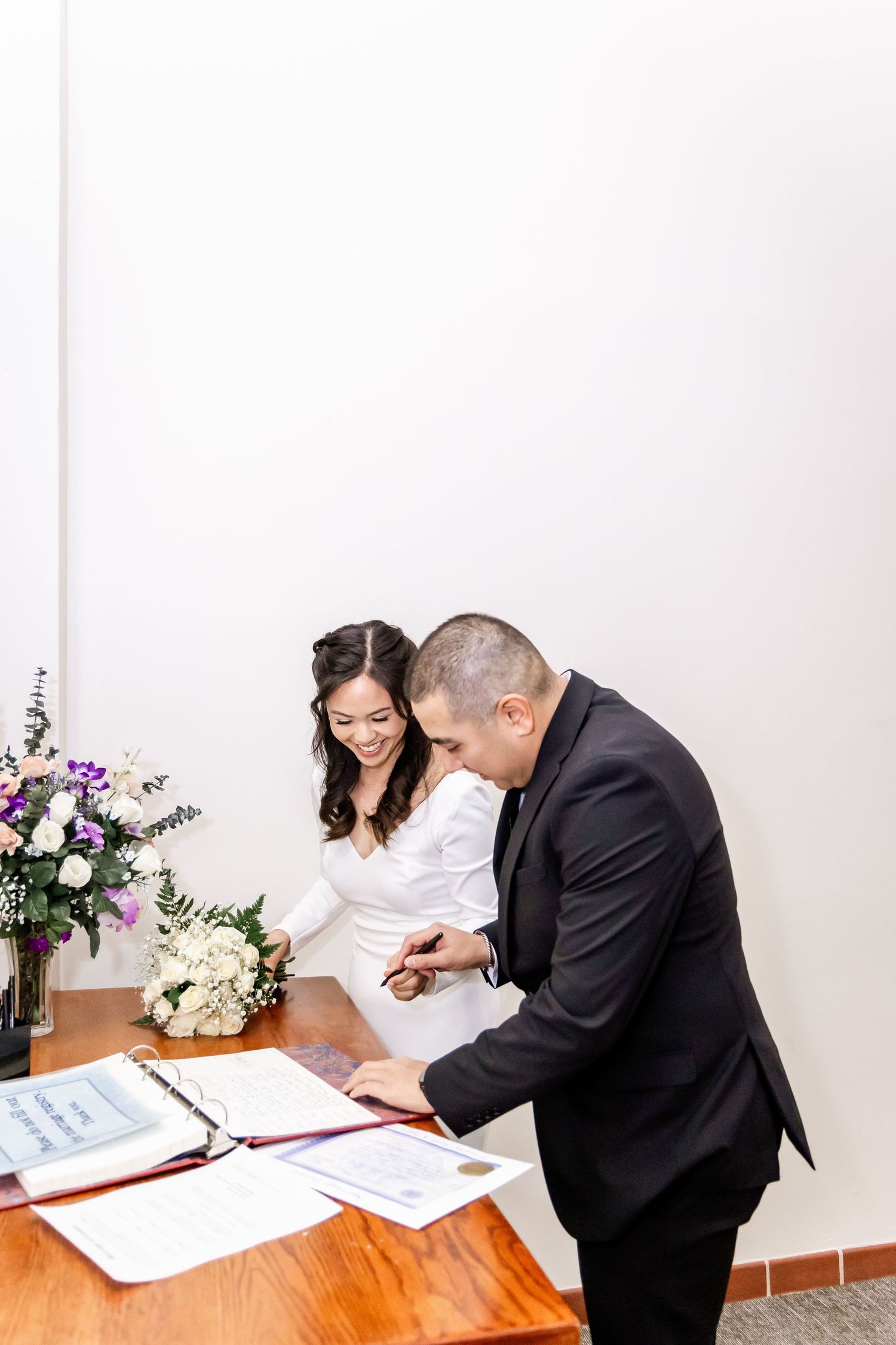Wedding couple signs marriage documents at a wooden table with floral arrangements.