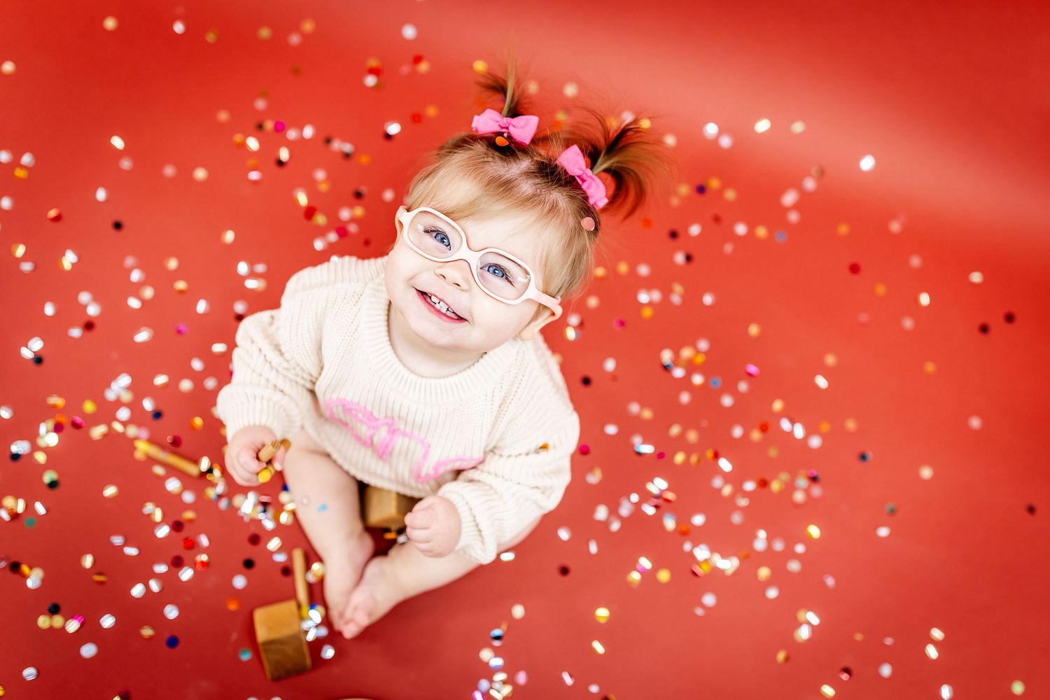 Happy baby wearing glasses and pink sweater plays with confetti on red background.