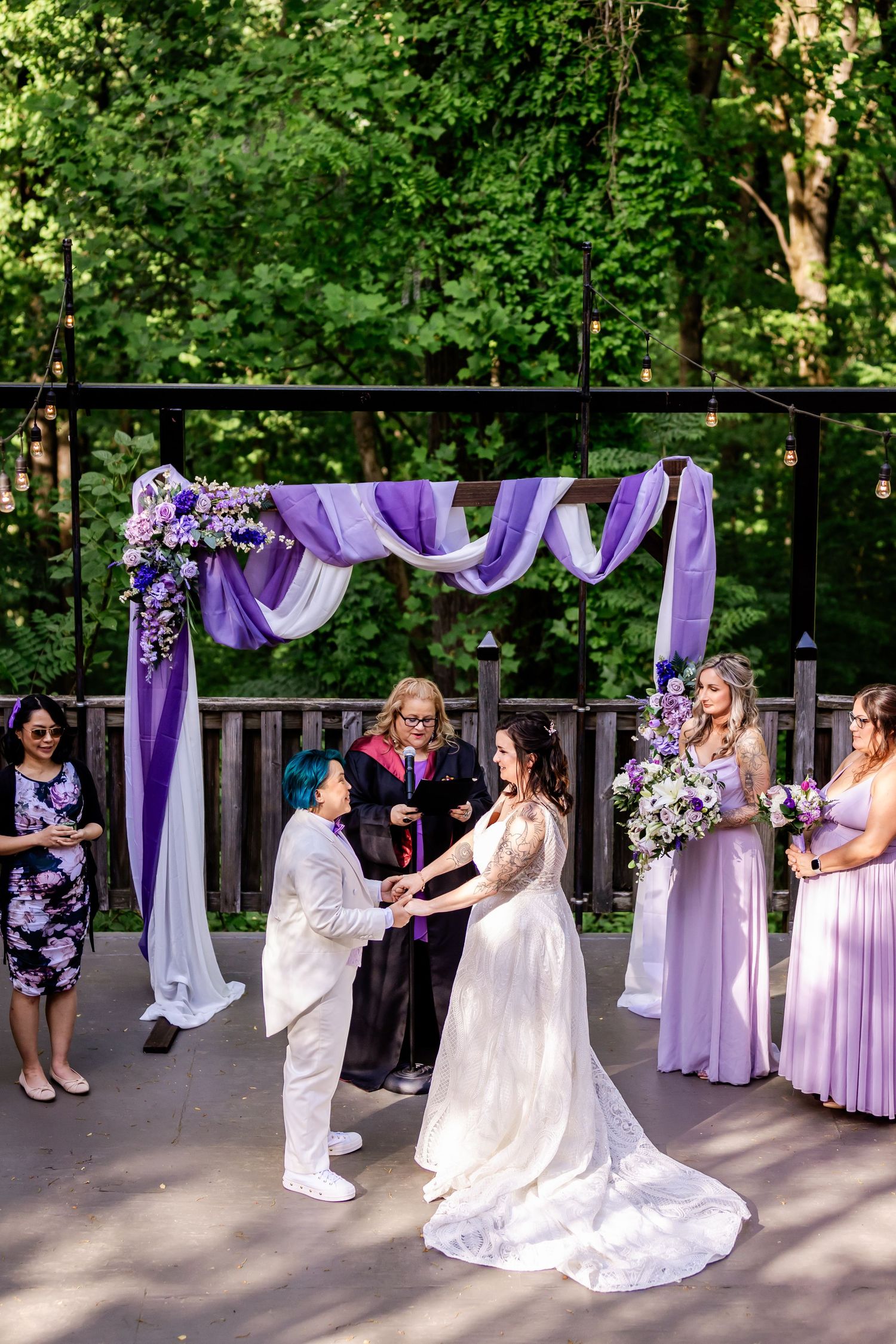 An outdoor wedding ceremony with purple draping, white flowers, and bridesmaids in lavender dresses.