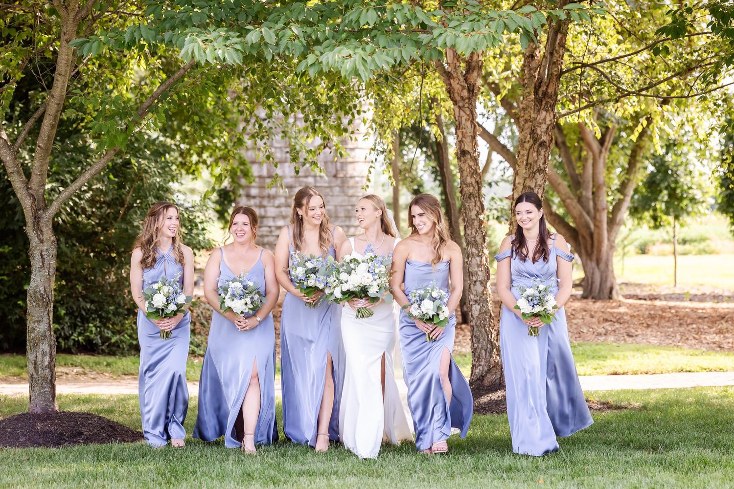 Bridesmaids wearing matching light blue gowns hold bouquets while standing together outdoors under leafy trees.