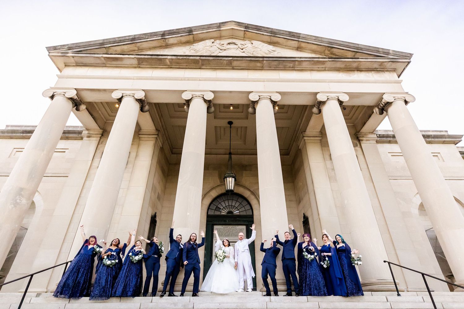 Wedding group celebrates with raised arms on historic building steps with tall white columns.