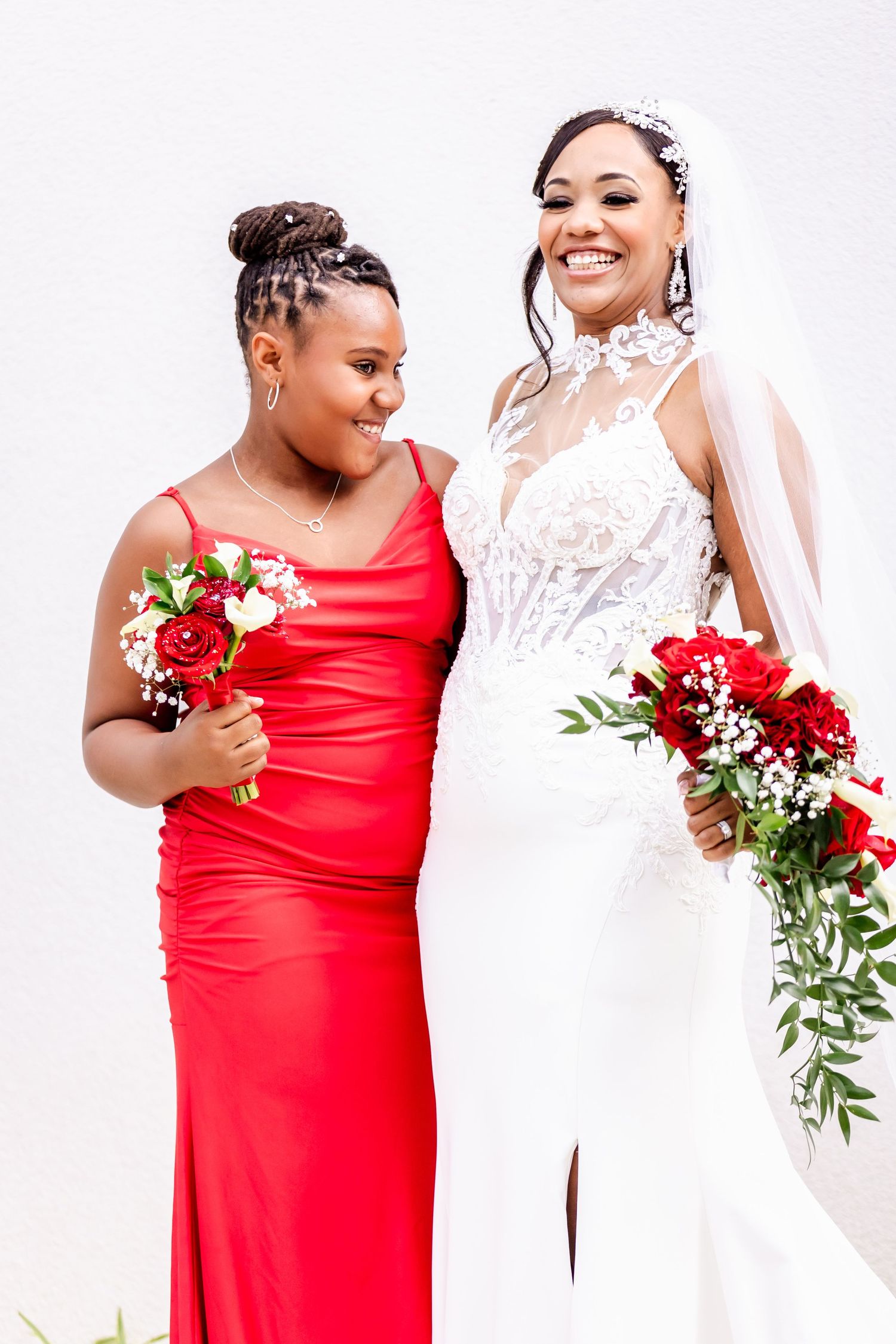 Two people pose together holding red and white floral bouquets against white background.