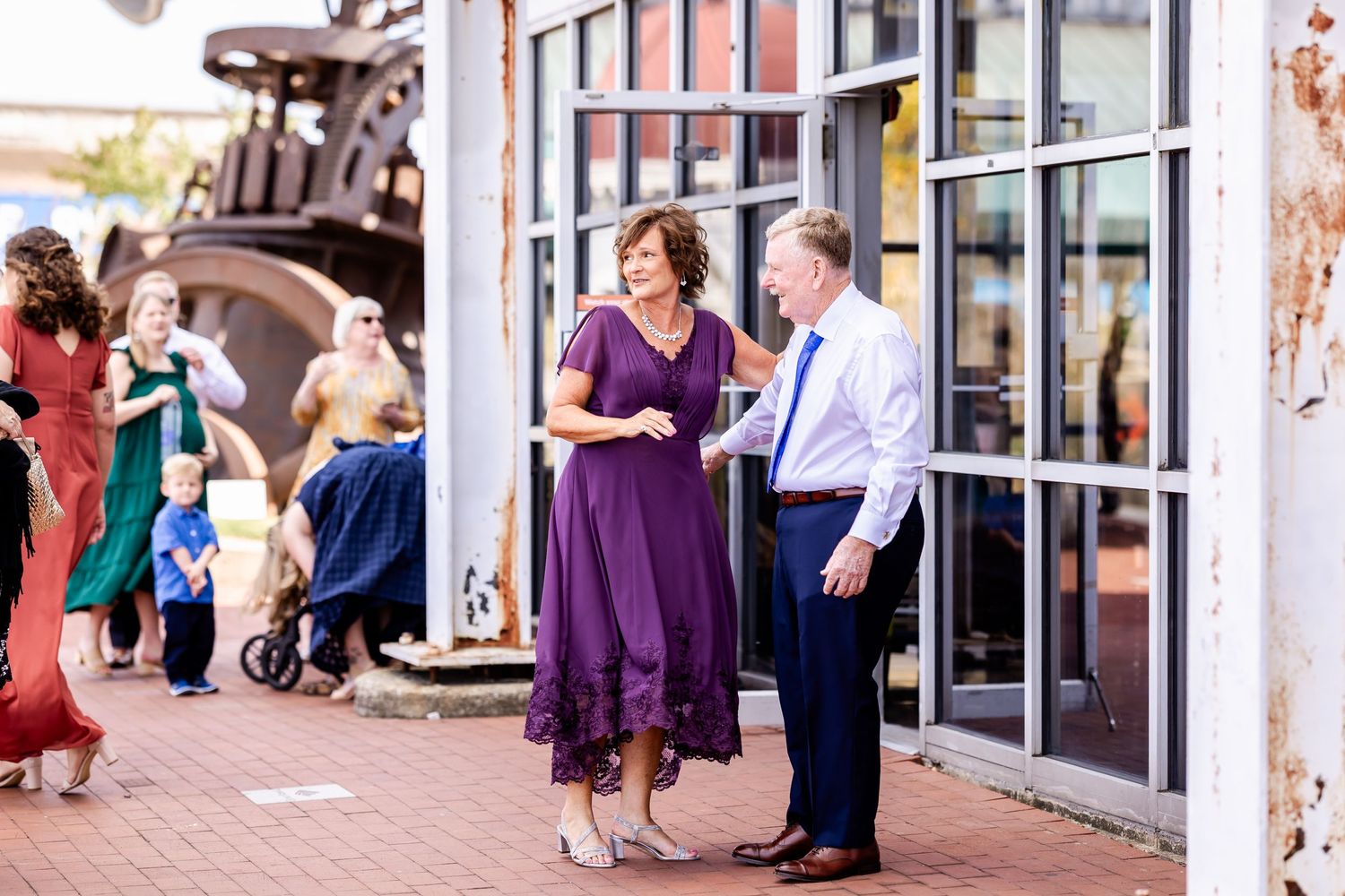 Wedding guests in formal attire gathered outside a brick building on a sunny day.