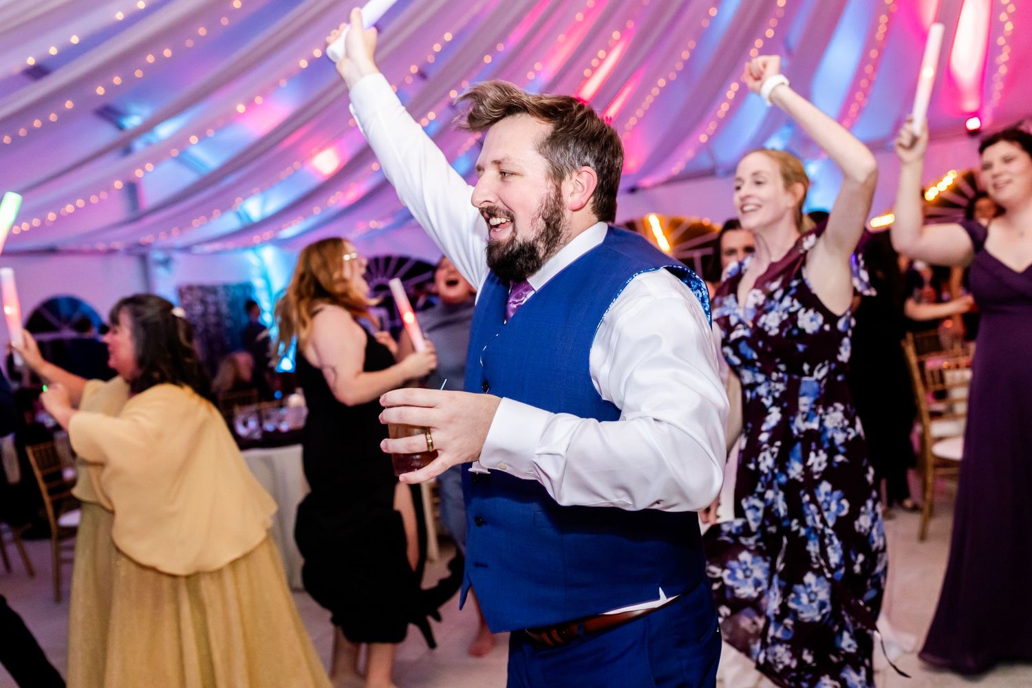 Celebration guest in blue vest enjoying lively dance moment under draped tent ceiling.