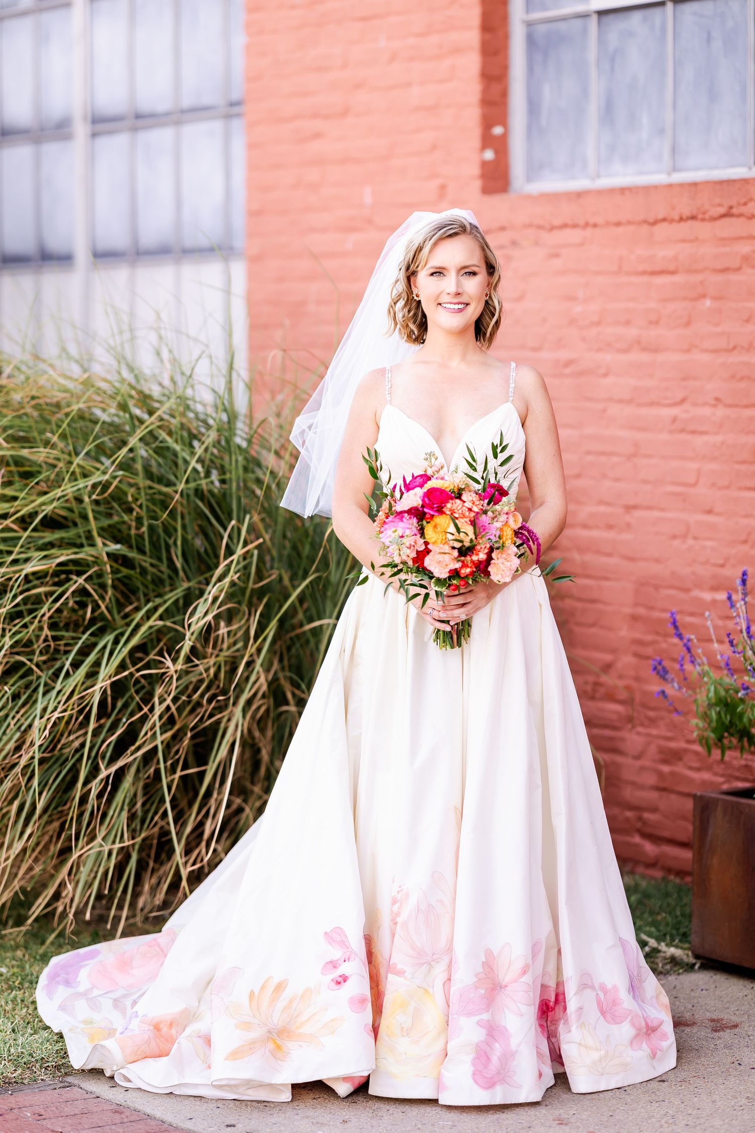 A bride in a white dress with pink floral details poses with a colorful bouquet against a rustic brick wall.