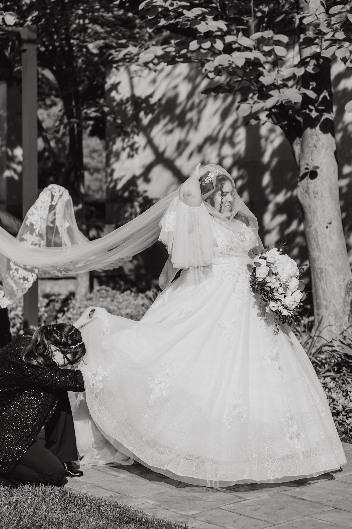 Black and white portrait of bride in flowing dress and veil holding bouquet in garden setting.
