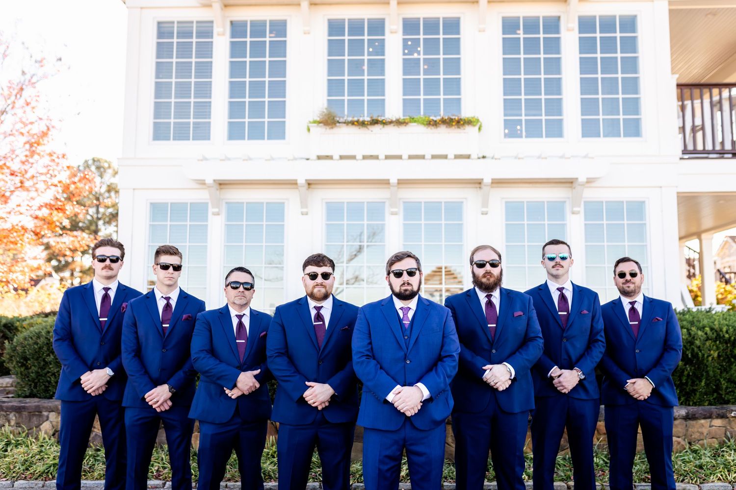 Nine men in coordinated navy suits and red ties pose formally in front of a white mansion.